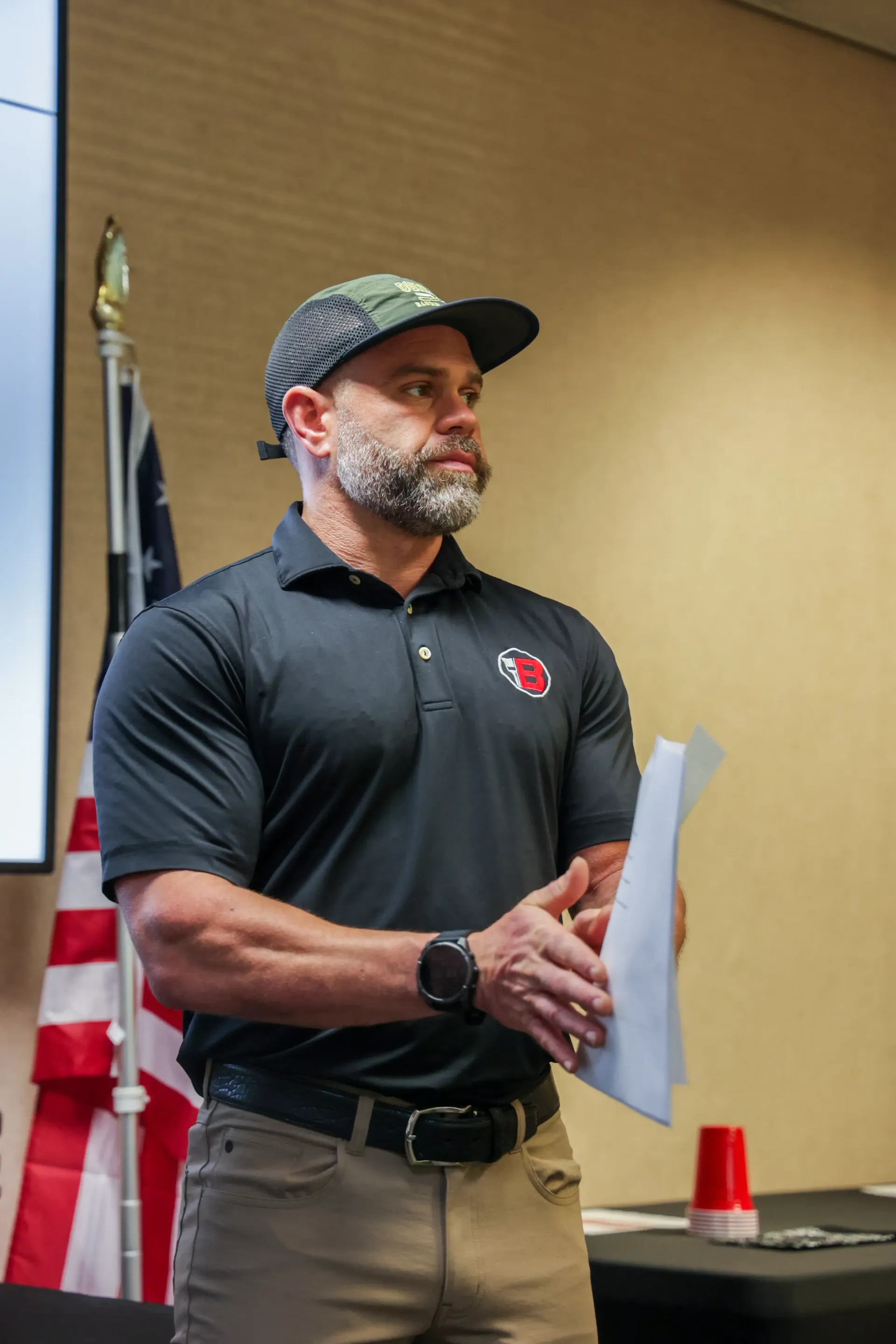 Man in black polo and camo cap holding papers and speaking at a podium indoors, with a U.S. flag nearby