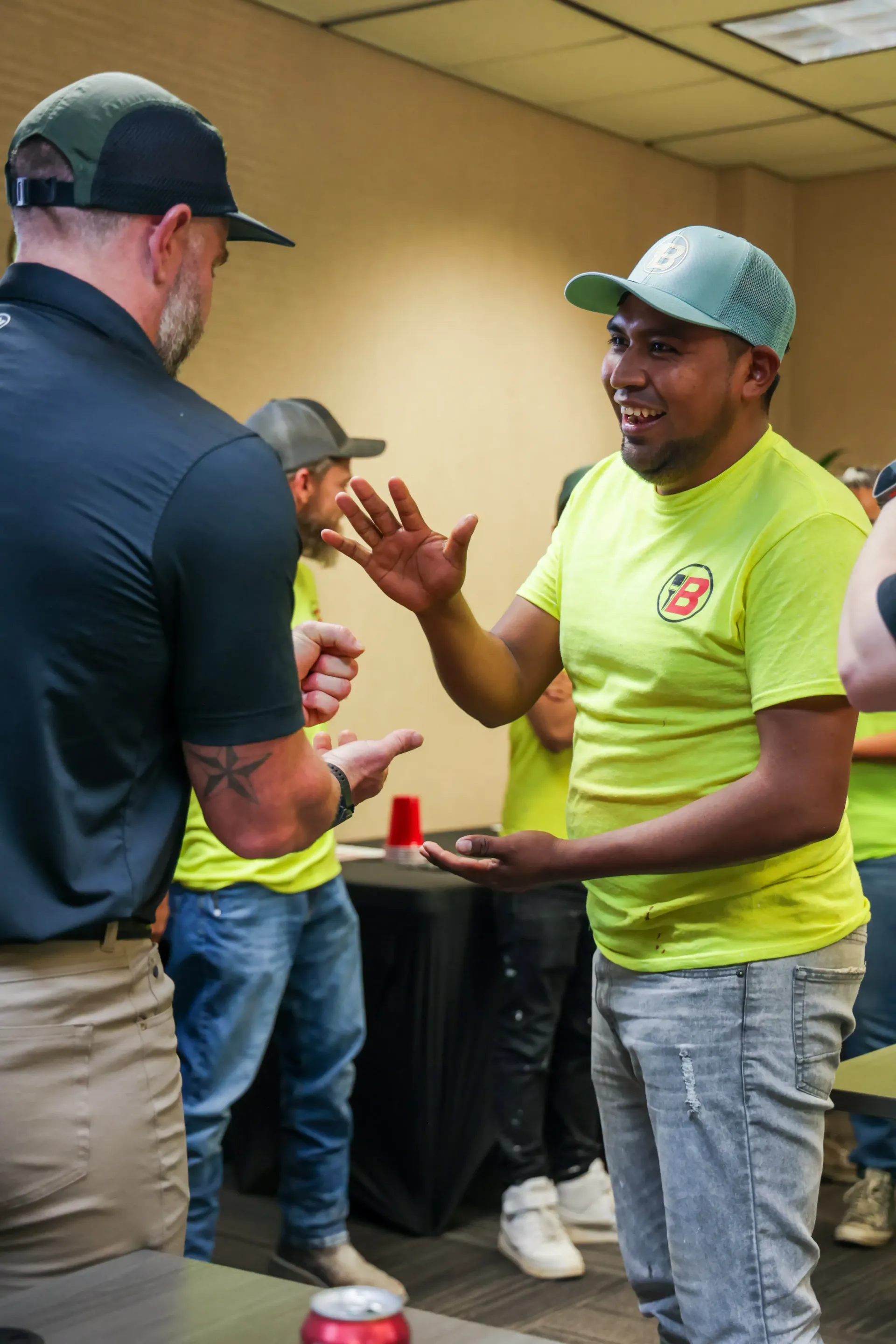 Two men in caps talking and gesturing in a room, one in a neon yellow shirt holding a tray.