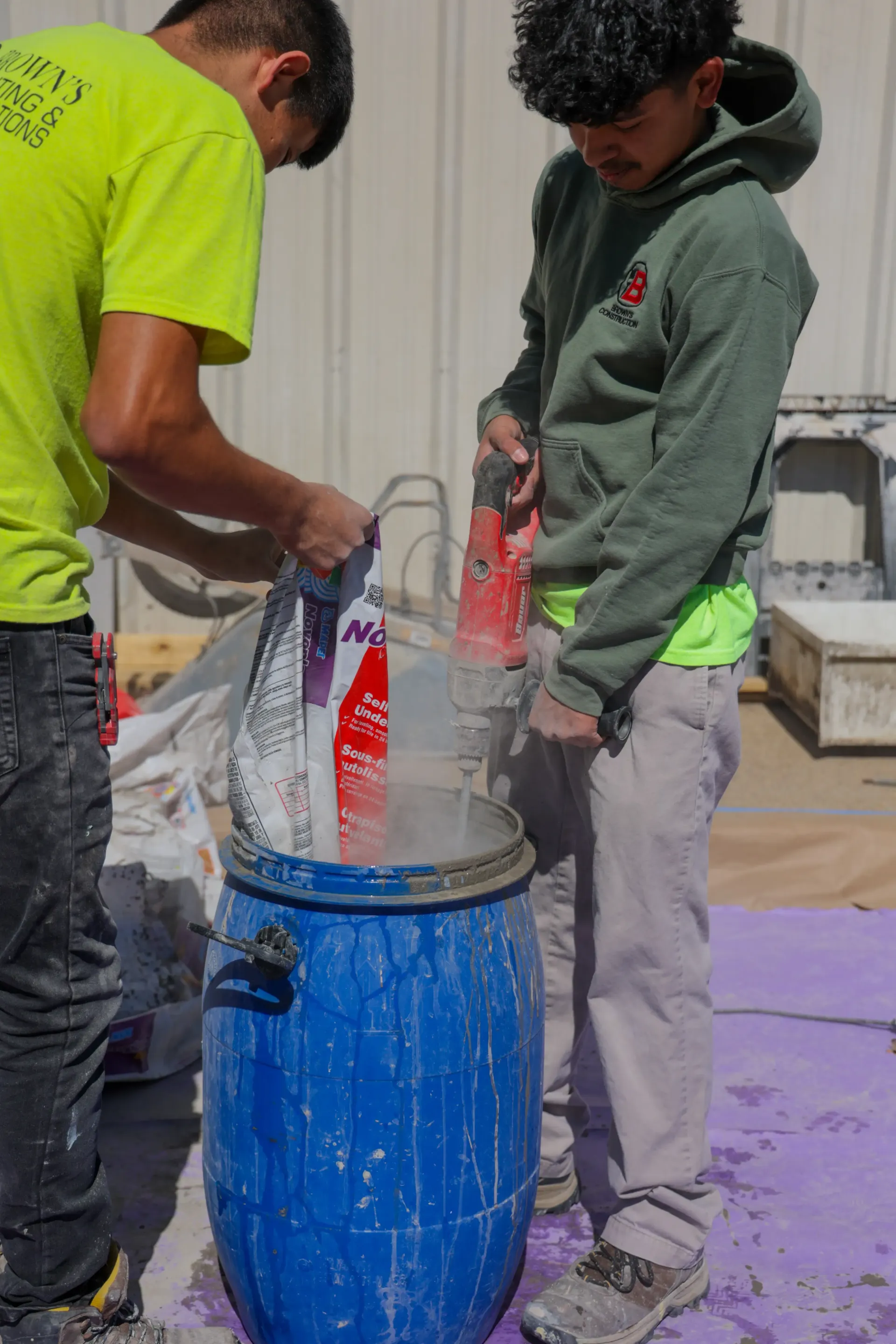 Two people pour material into a blue barrel in a workshop, with dust or powder visible around them.