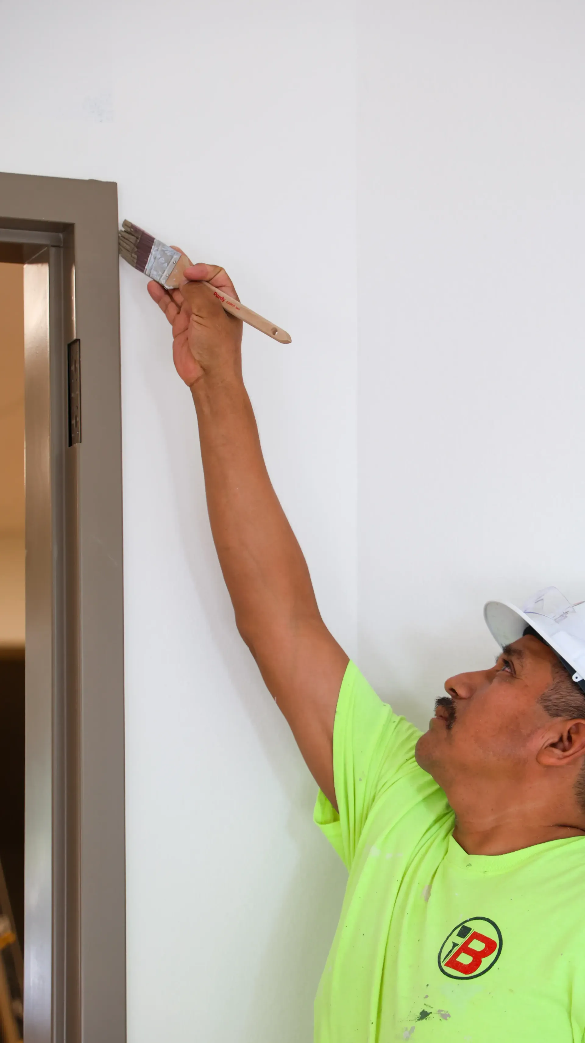 Worker painting a white wall with a brush, reaching up beside a doorway