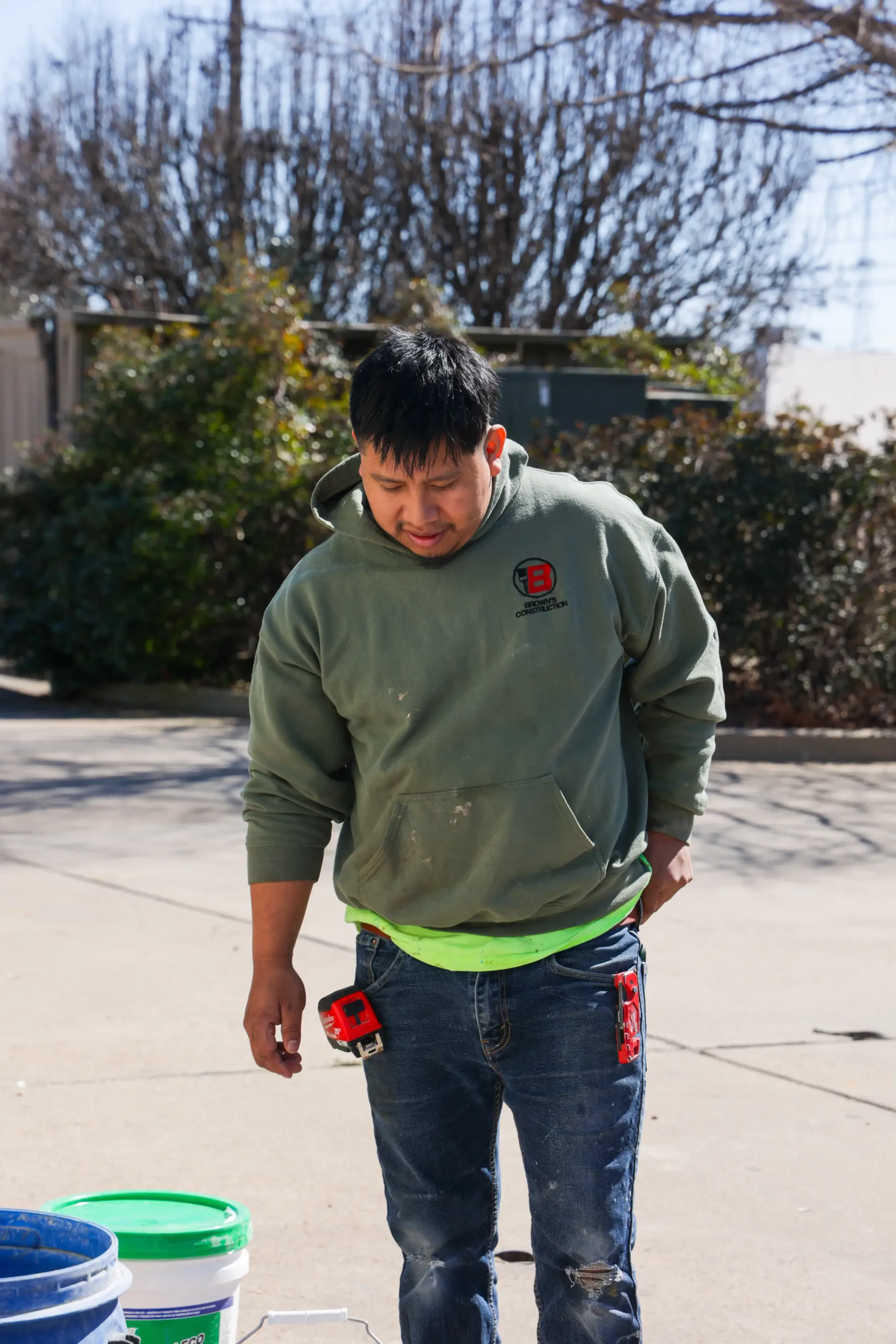 Person standing outdoors in a driveway, looking down, wearing a green sweatshirt and jeans.