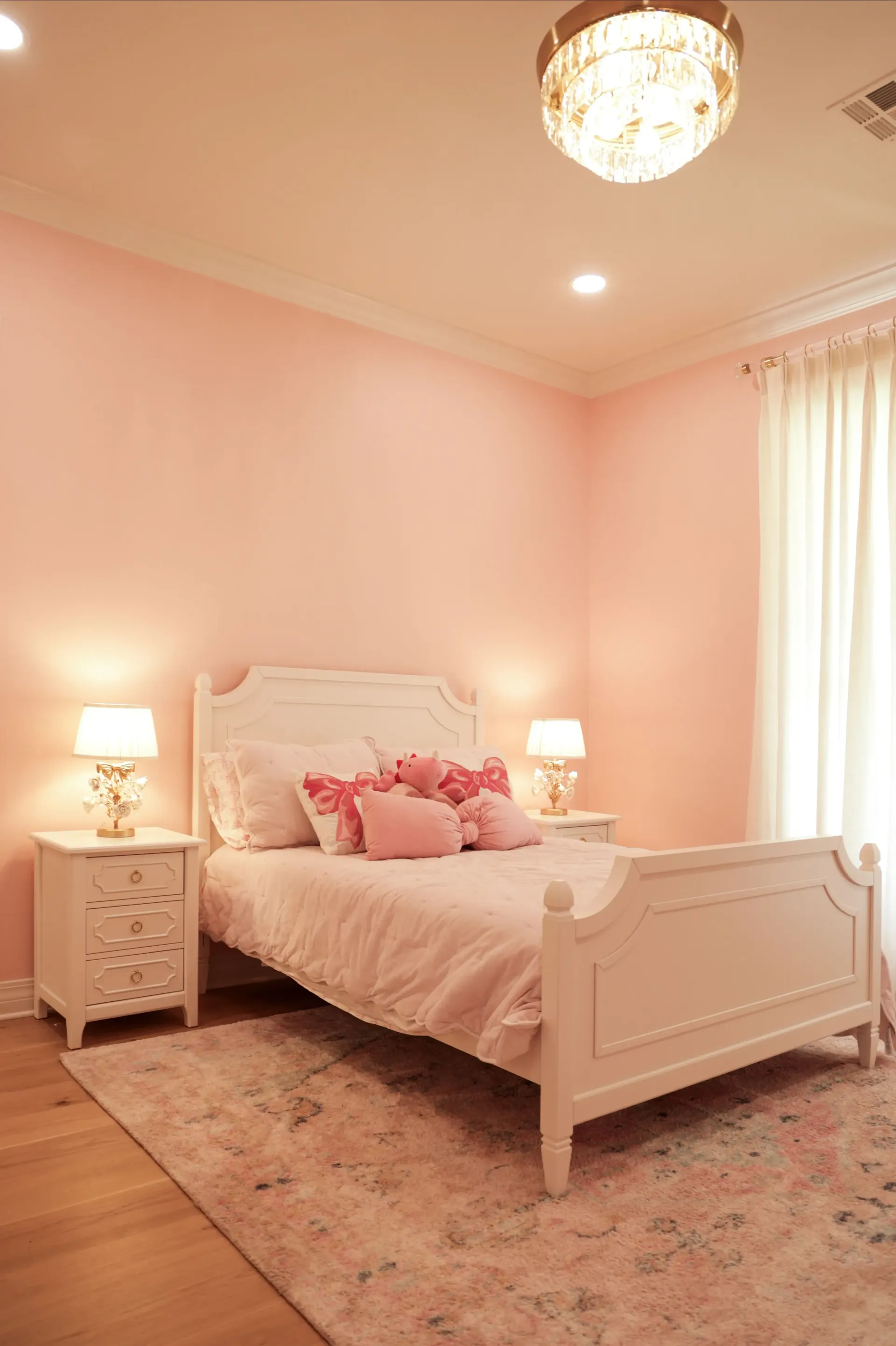Pink bedroom with a white bed, matching nightstands, lamps, rug, and a window with sheer curtains