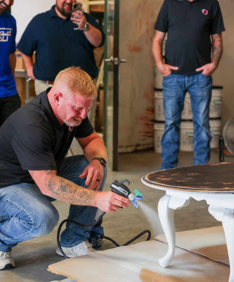 Man spray-painting a white table leg indoors while others watch nearby