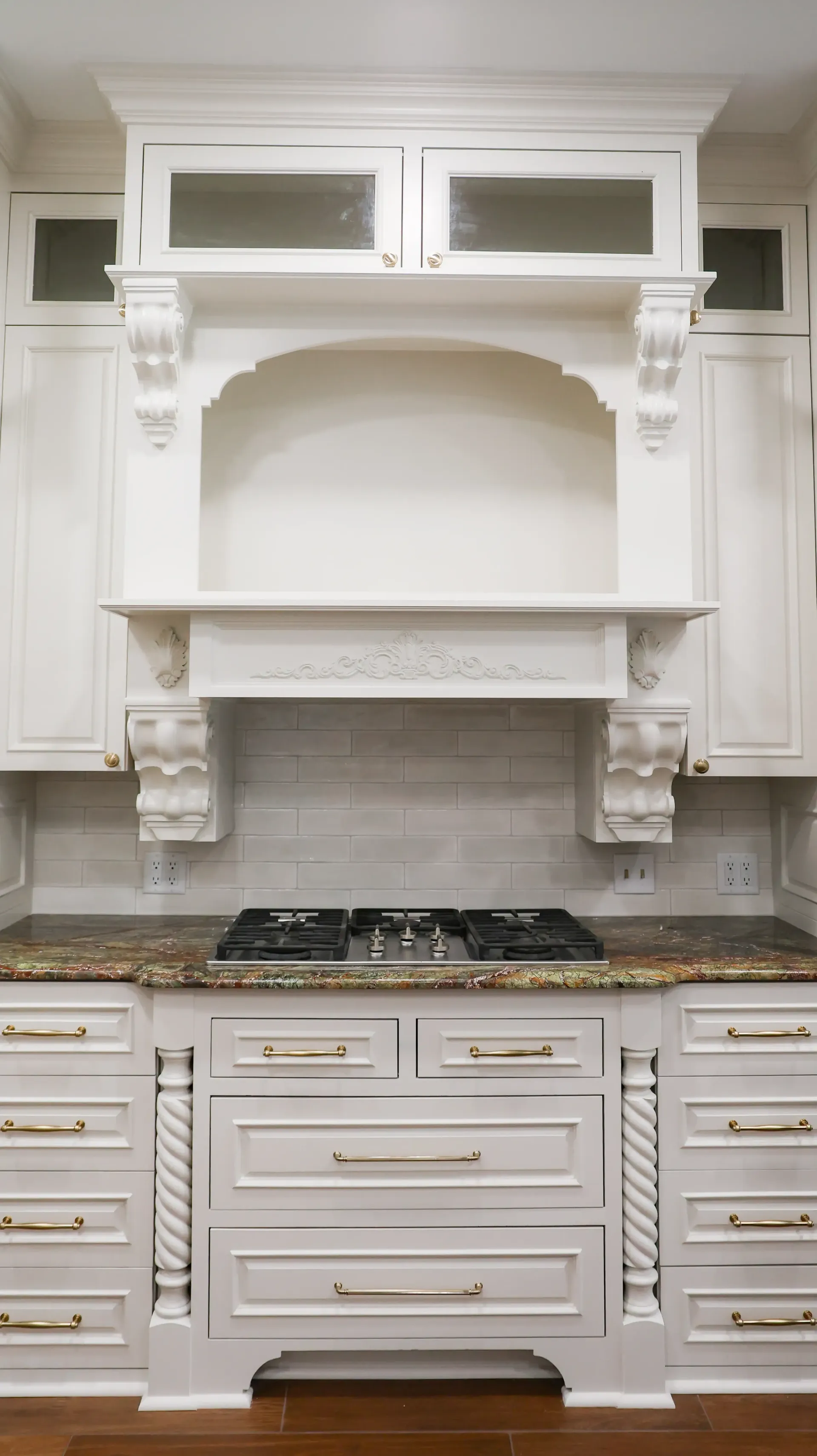 White kitchen with ornate range hood, cabinets, granite countertops, and built-in stovetop.