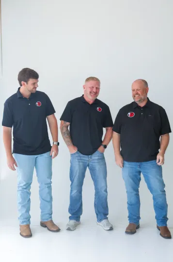 Three men in black polo shirts and jeans standing against a plain white wall.