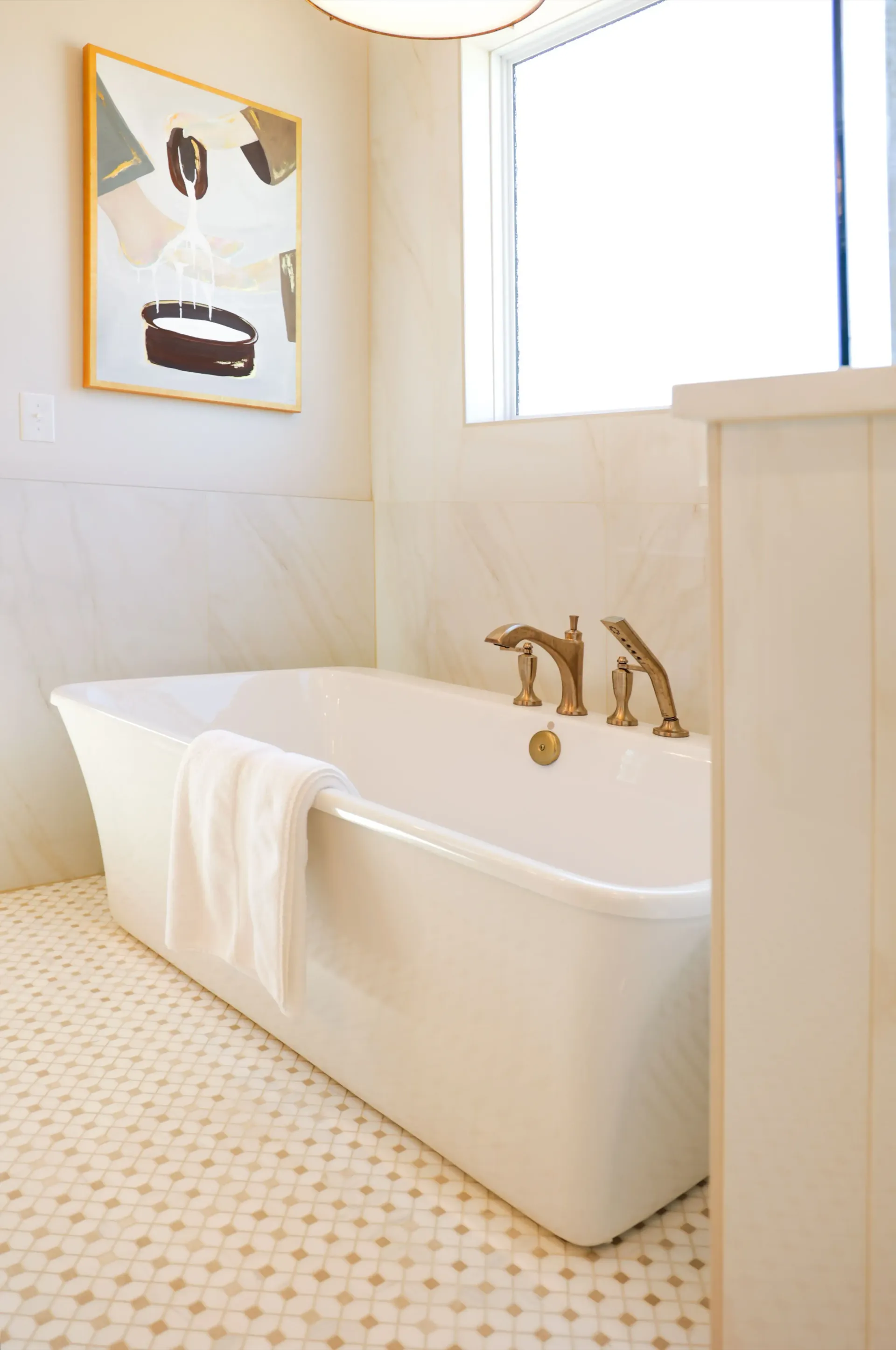 Bright white bathroom with a freestanding tub, gold faucet, window, and framed wall art.