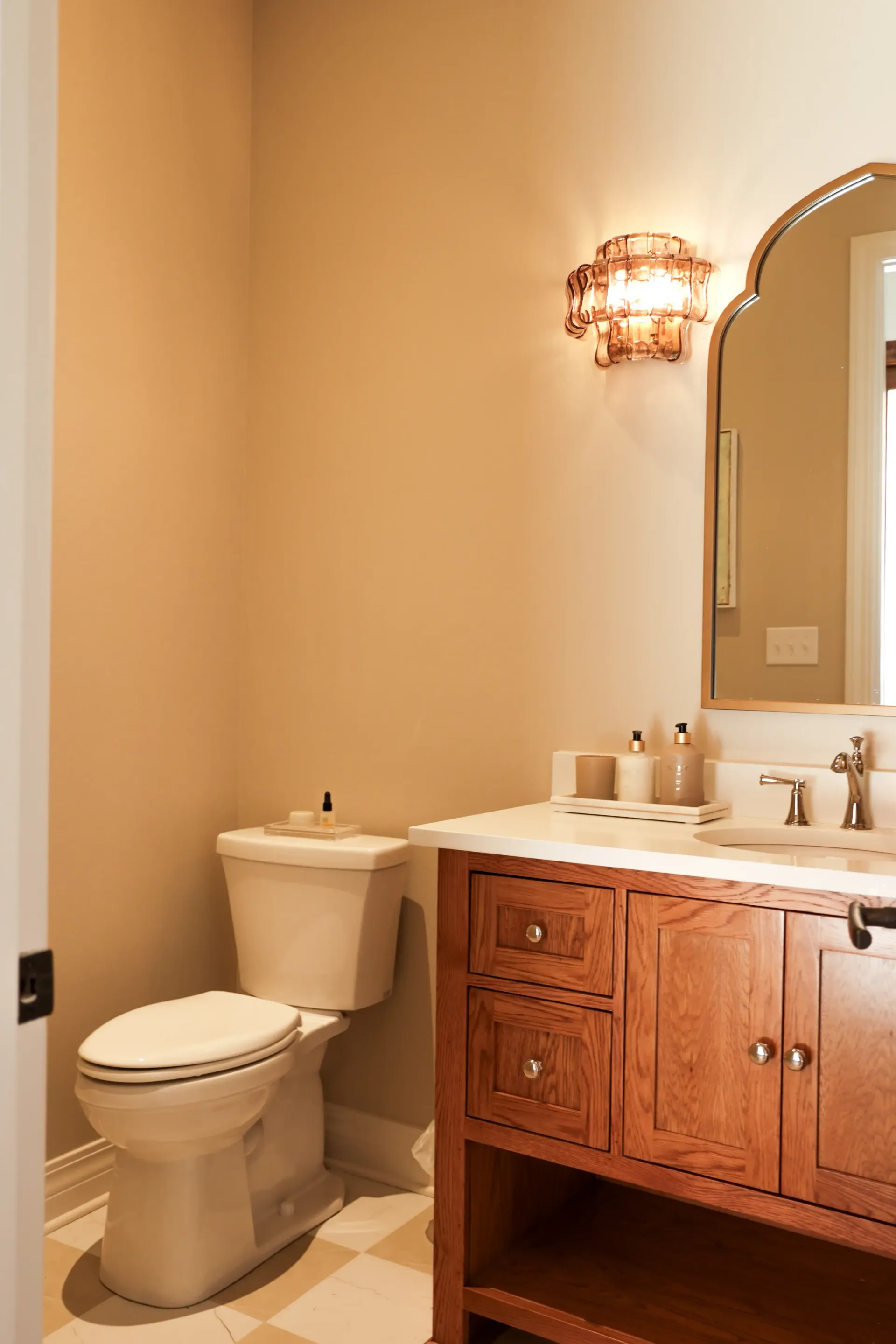 Small bathroom with toilet, wooden vanity, mirror, and wall sconce in warm beige tones