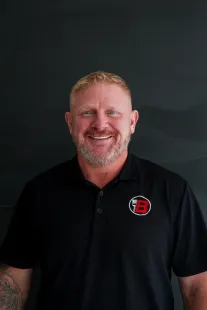 Smiling man in black polo shirt with red-and-white logo against dark background