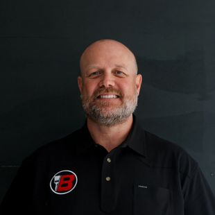 Smiling bald man in black polo with red-and-white logo, standing against a dark background