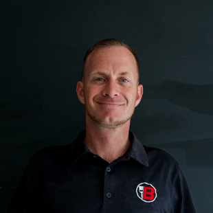 Portrait of a smiling man in a black polo with a red “B” logo against a dark background
