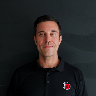 Portrait of a man in a black polo shirt with a red-and-white logo against a dark background