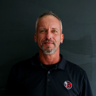 Portrait of a man in a black polo shirt with a red-and-white logo against a dark background