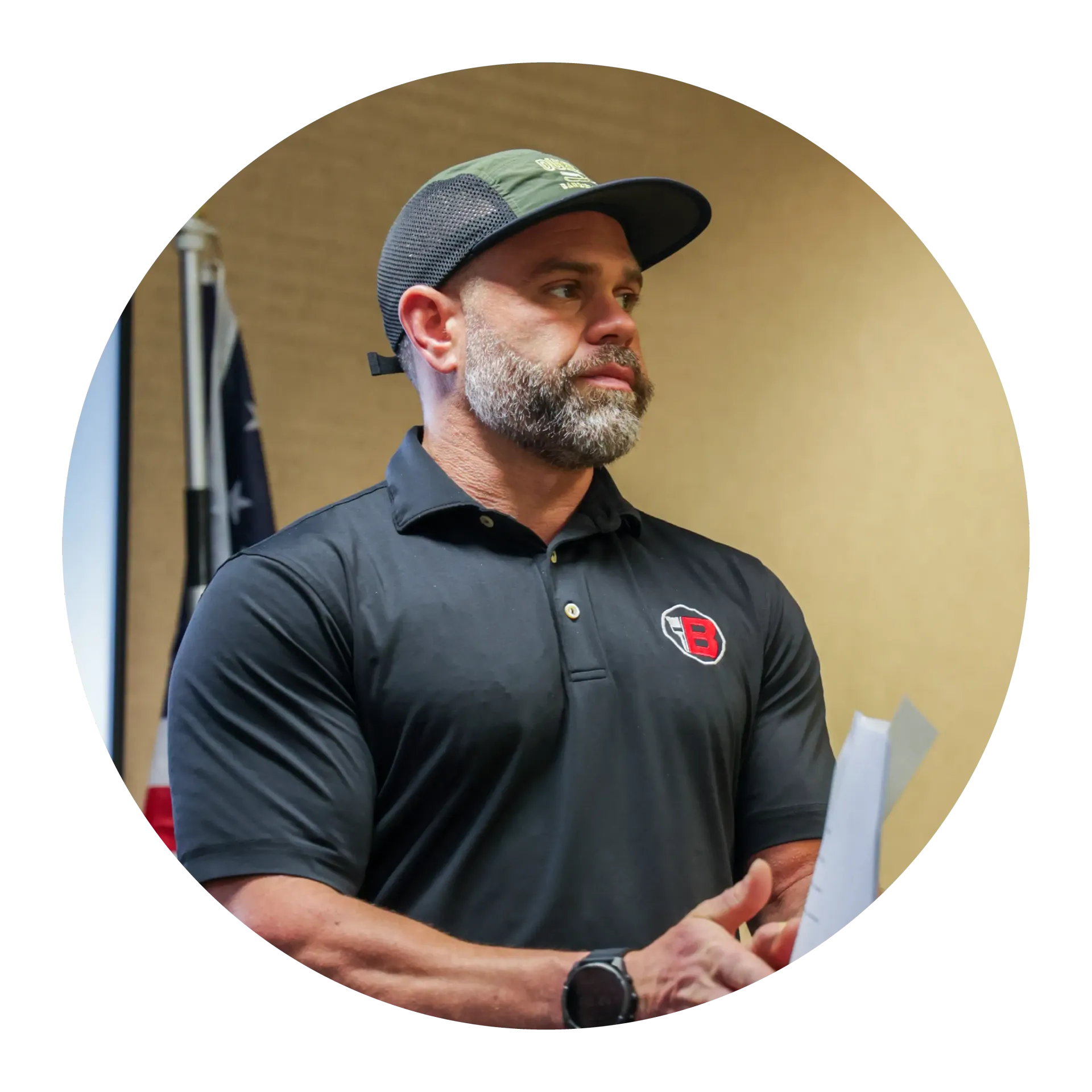 Man in black polo and cap speaking indoors, holding papers in a meeting room.