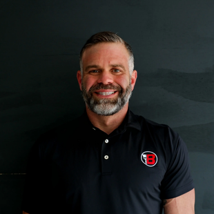 Smiling man in a black polo with a red-and-white logo, standing against a dark background