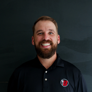 Smiling bearded man in a black polo with a red-and-white logo on a dark background