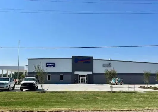 Wide industrial building with a white facade, blue signage, and parked vehicles under a clear sky