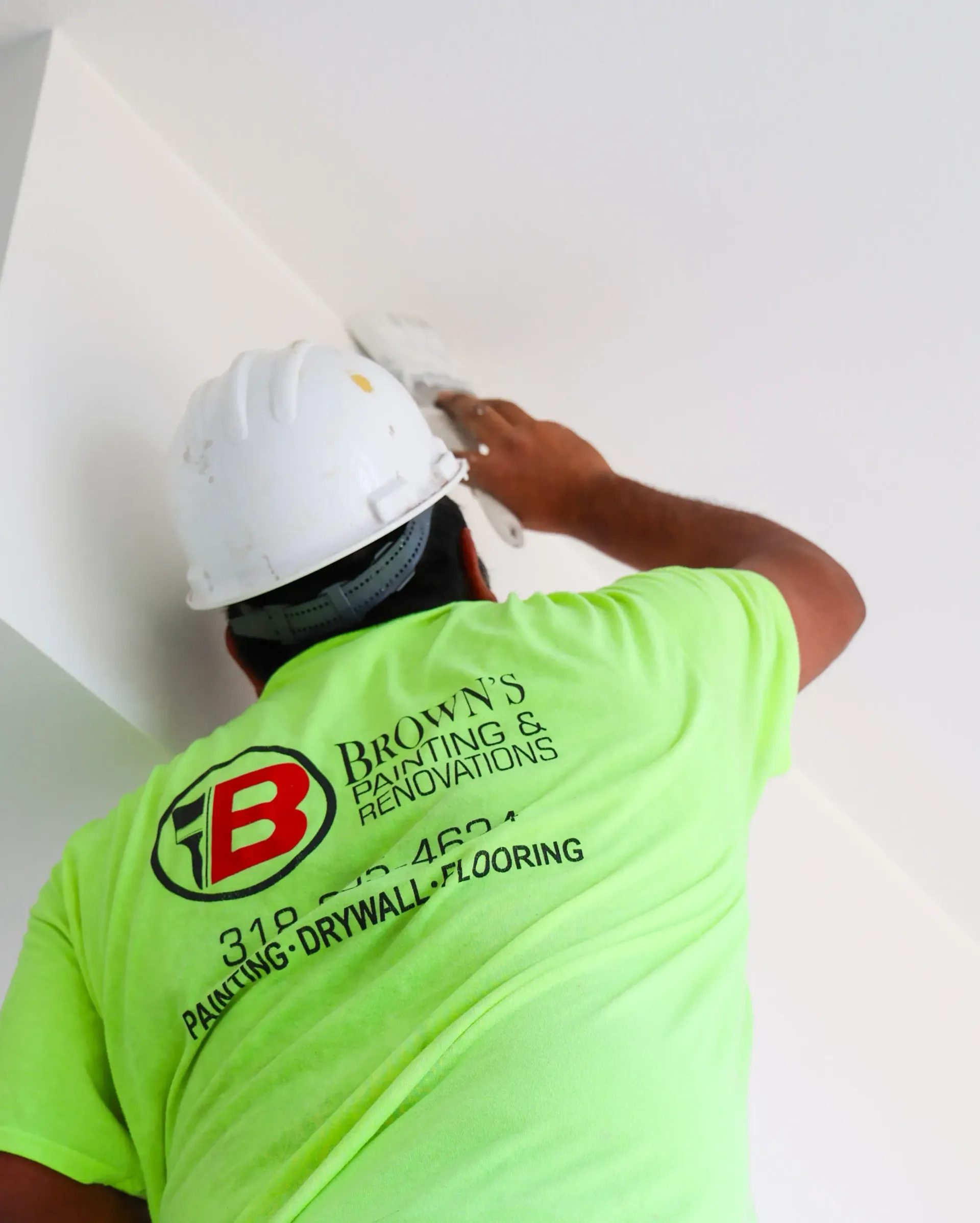 Worker in a neon green shirt and white hard hat painting a white ceiling.