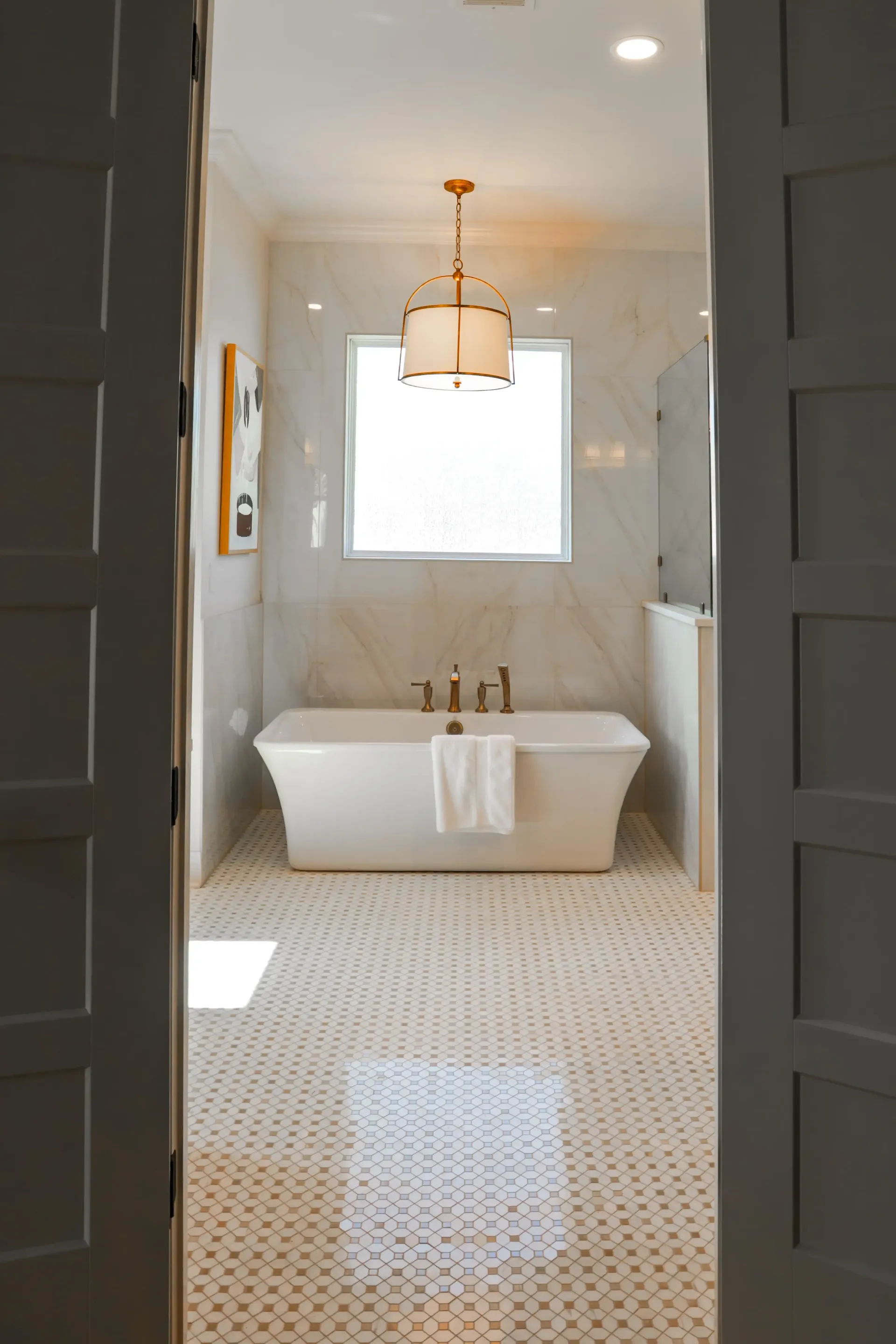 Bright bathroom with a freestanding tub, gold chandelier, and patterned tile floor, viewed through open doors