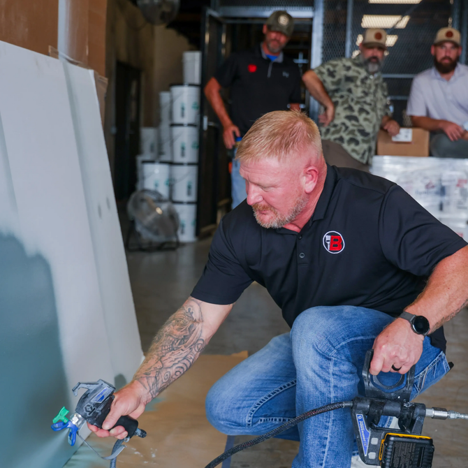 Man crouching with a power tool in a workshop, with two men standing in the background.