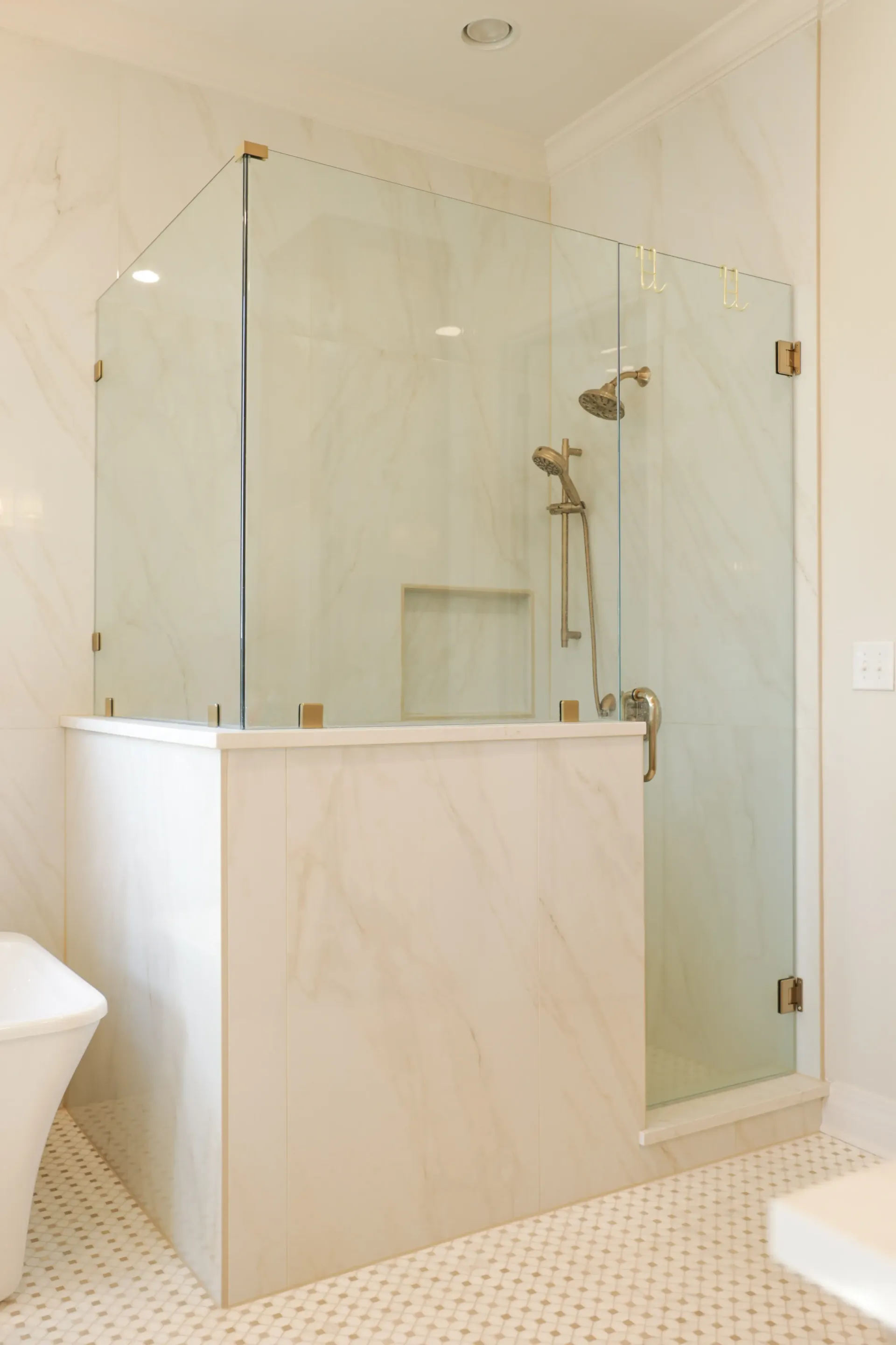 Glass-enclosed walk-in shower with marble half wall and beige tile in a bright bathroom