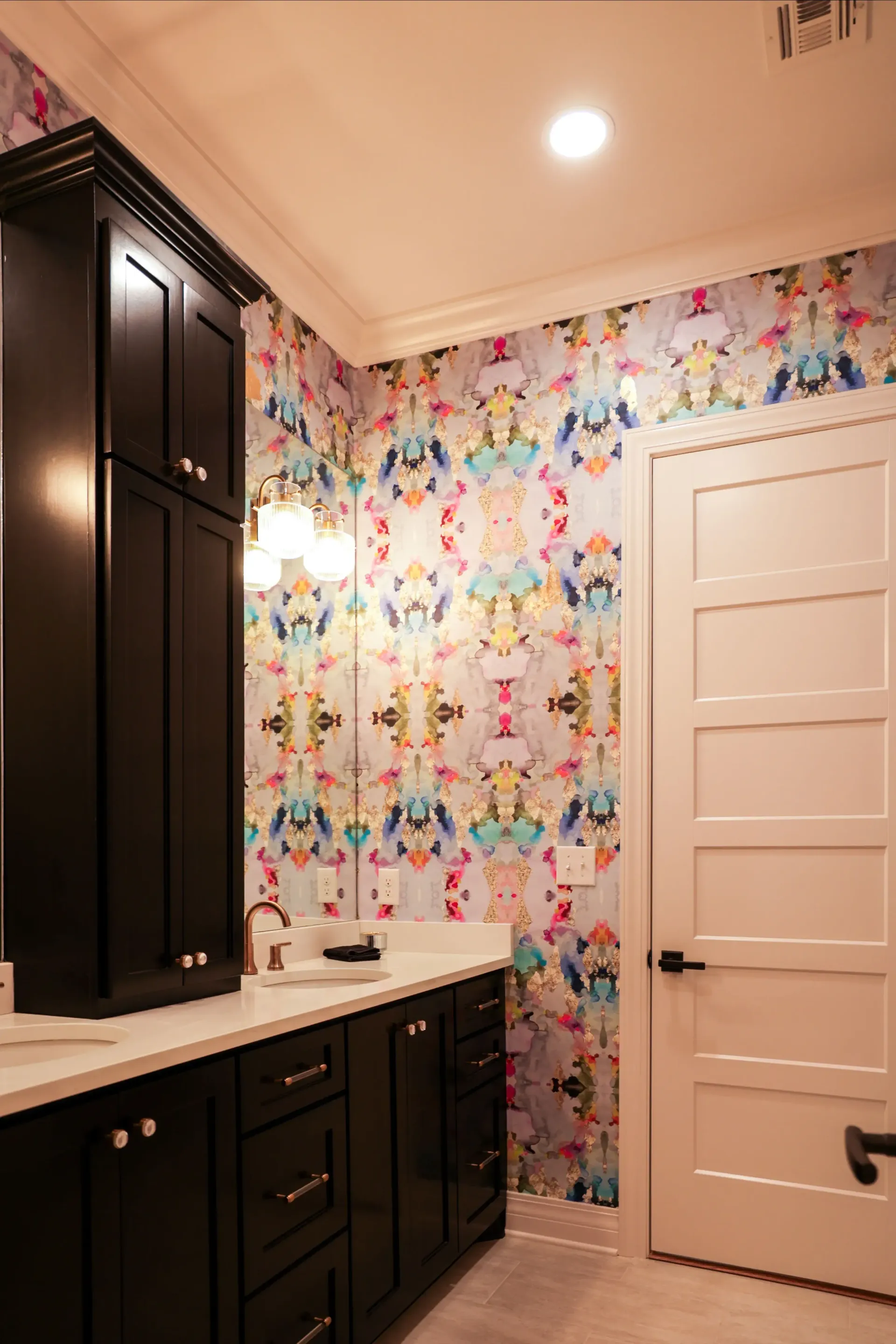 Bright bathroom with floral wallpaper, dark cabinets, white countertops, and a white door.