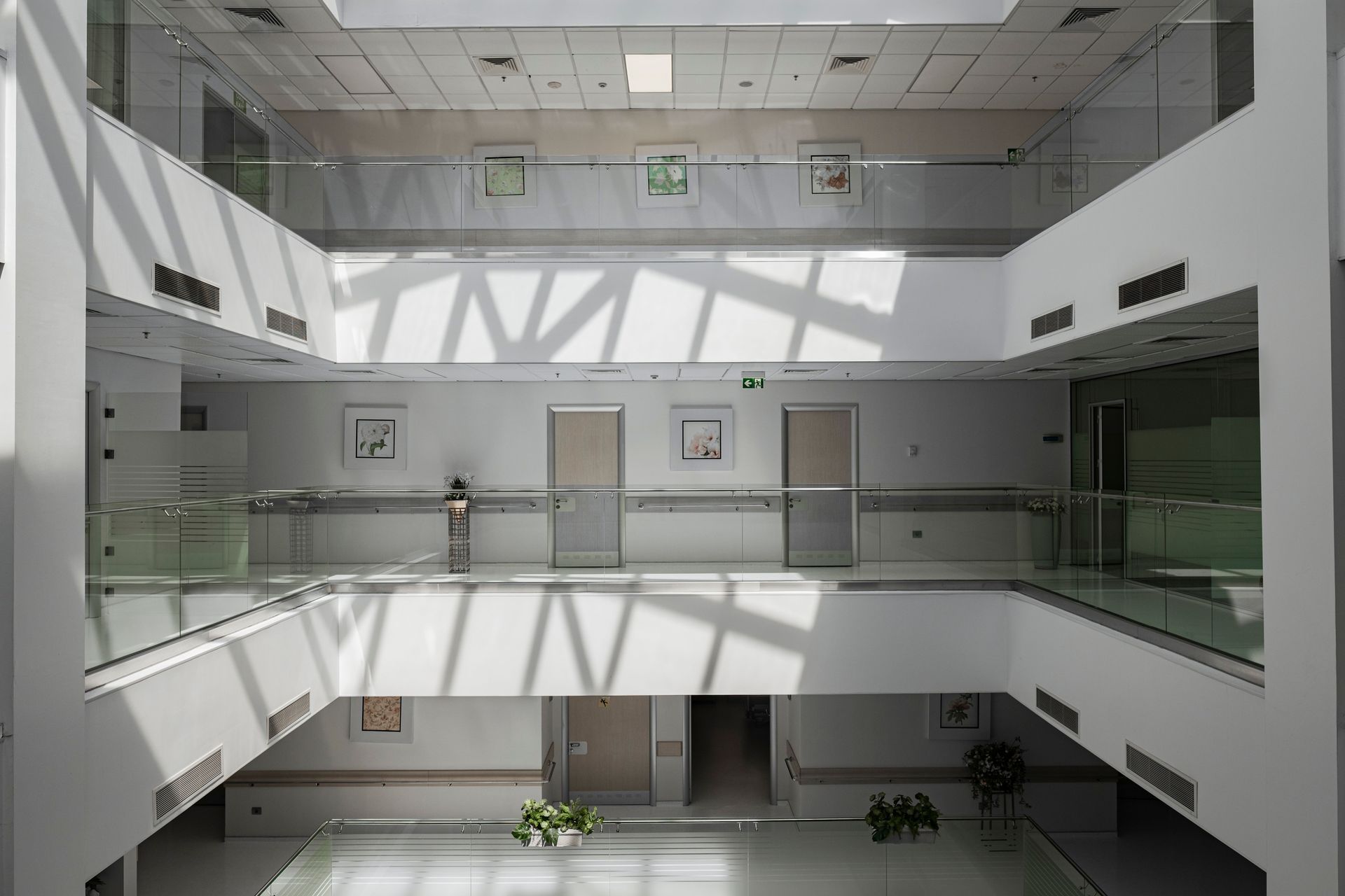Bright atrium with glass railings, white walls, skylights, and sunlight casting geometric shadows.