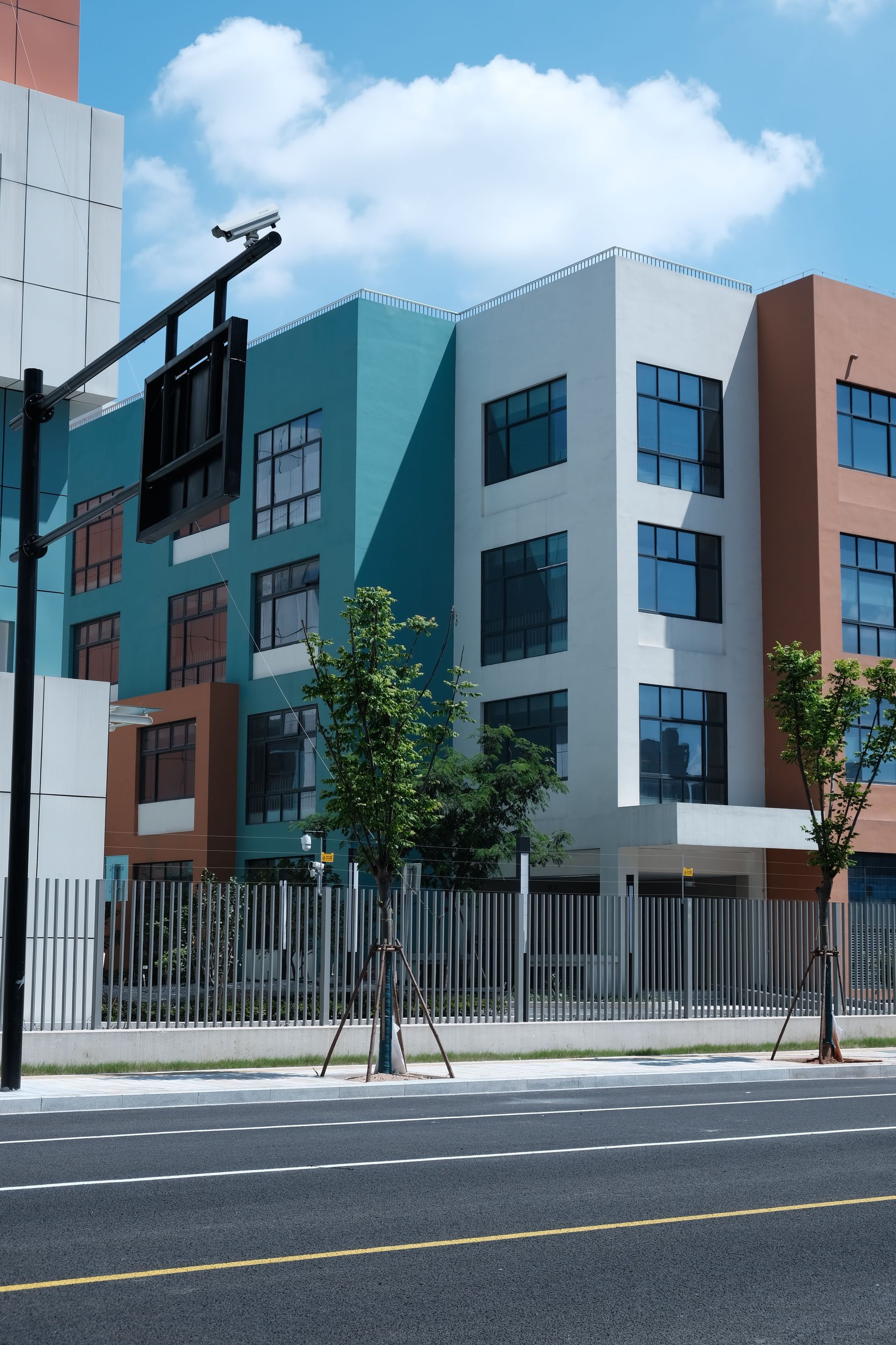 Modern multi-story apartment building with teal, white, and brown facades behind a street and fence