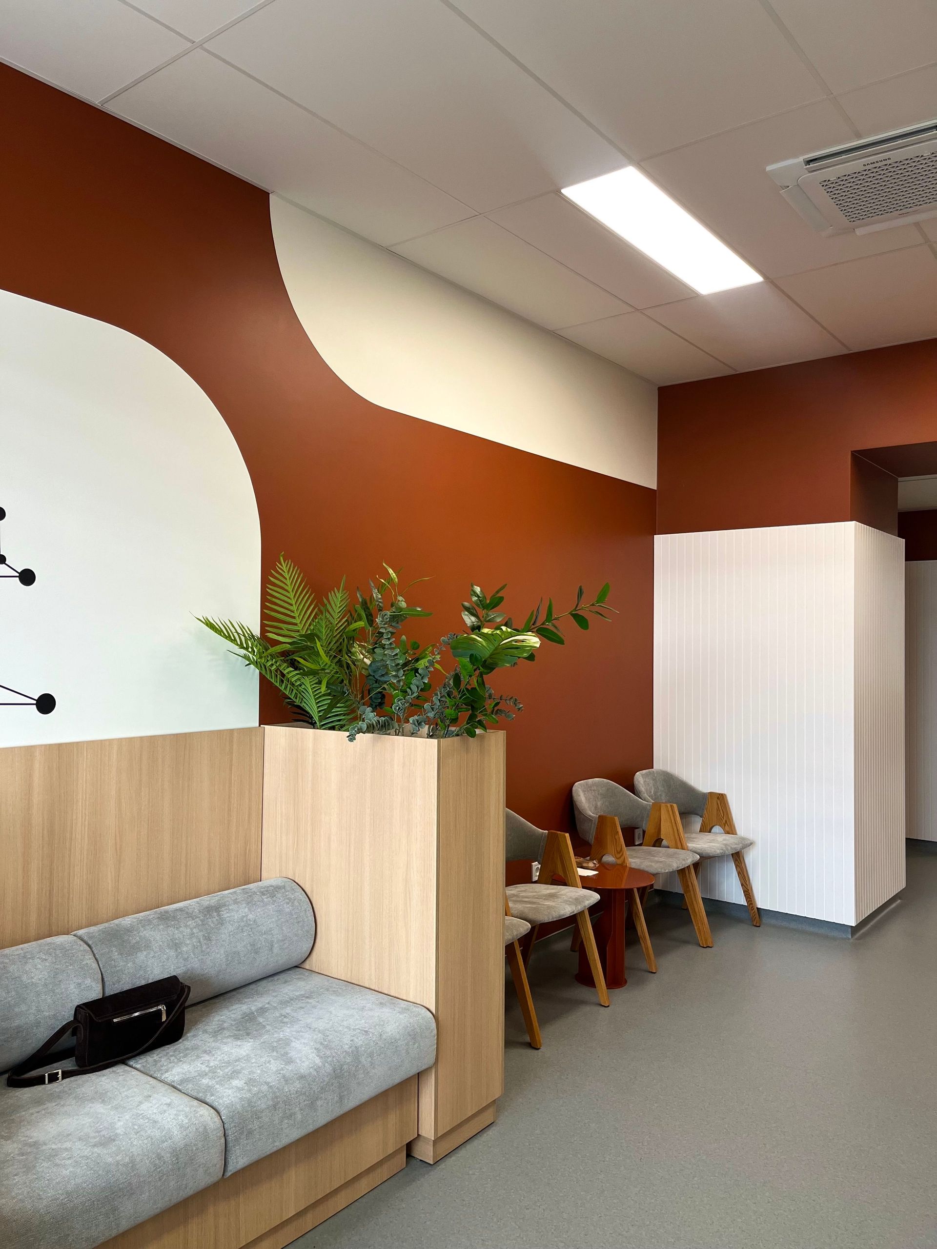 Modern office lounge with gray couch, plant divider, chairs, and a white storage cabinet against red walls.
