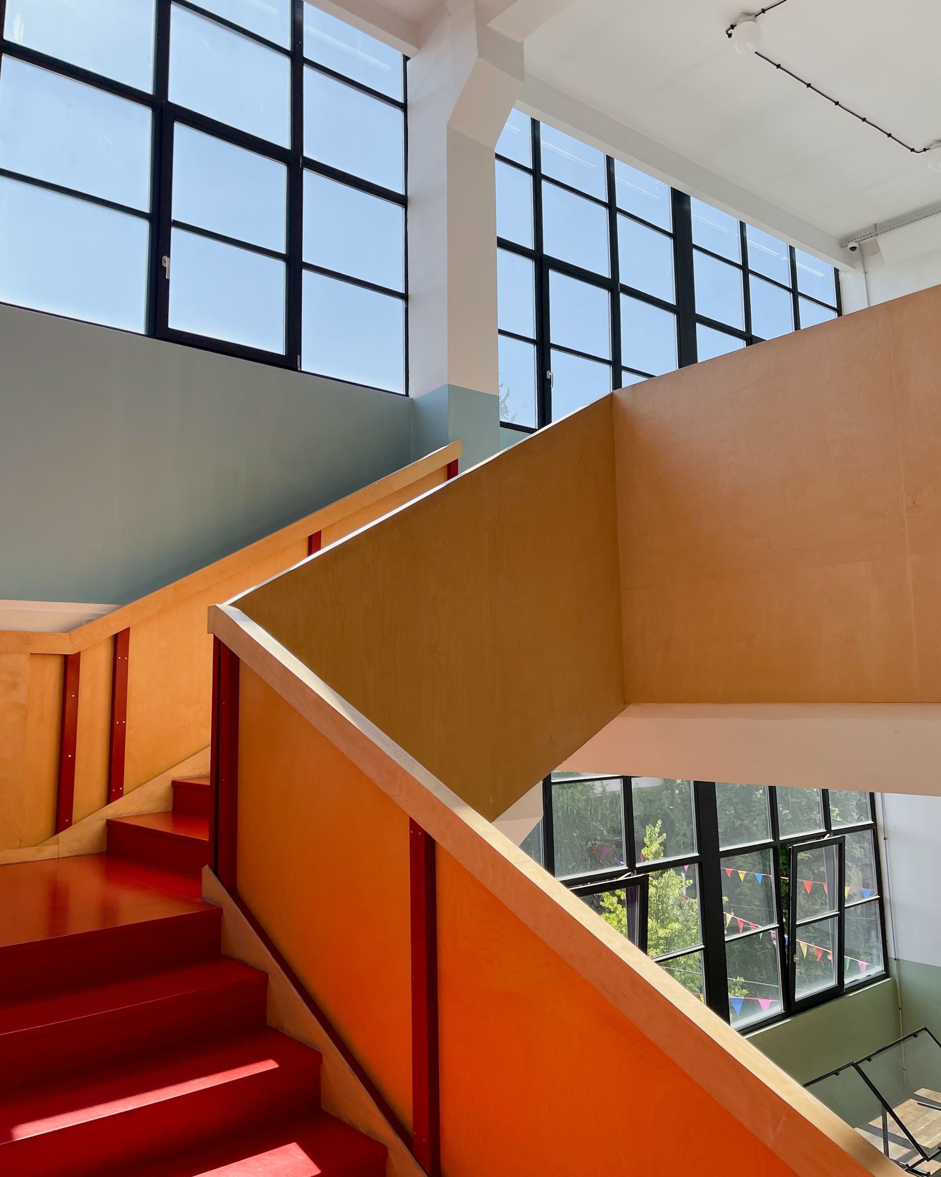Bright indoor stairwell with red steps, orange railings, and large windows overlooking trees.