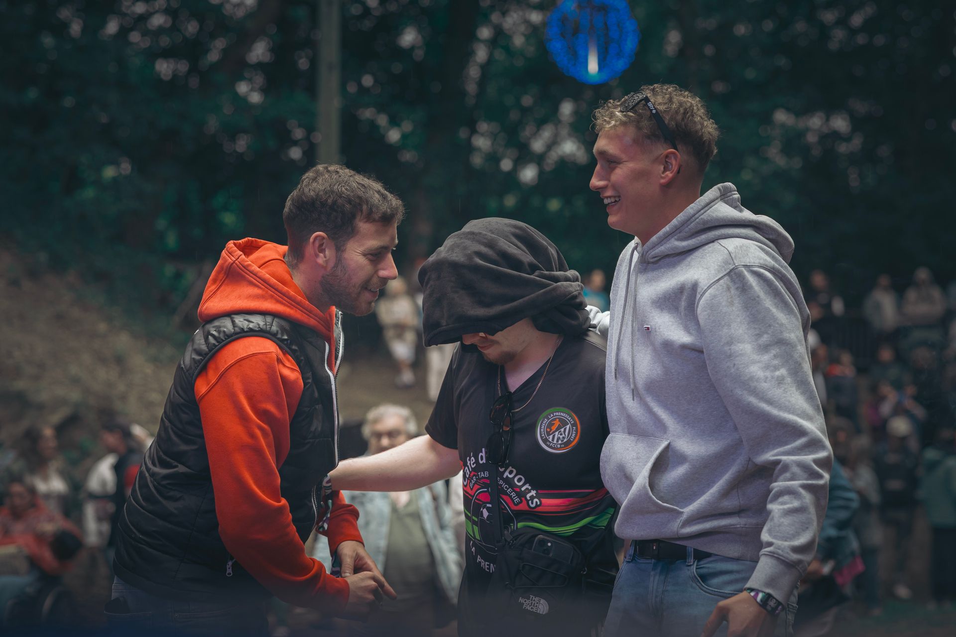 Trois personnes discutent dehors, l'une porte un gilet orange, l'autre une casquette noire et la dernière un sweat à capuche gris.