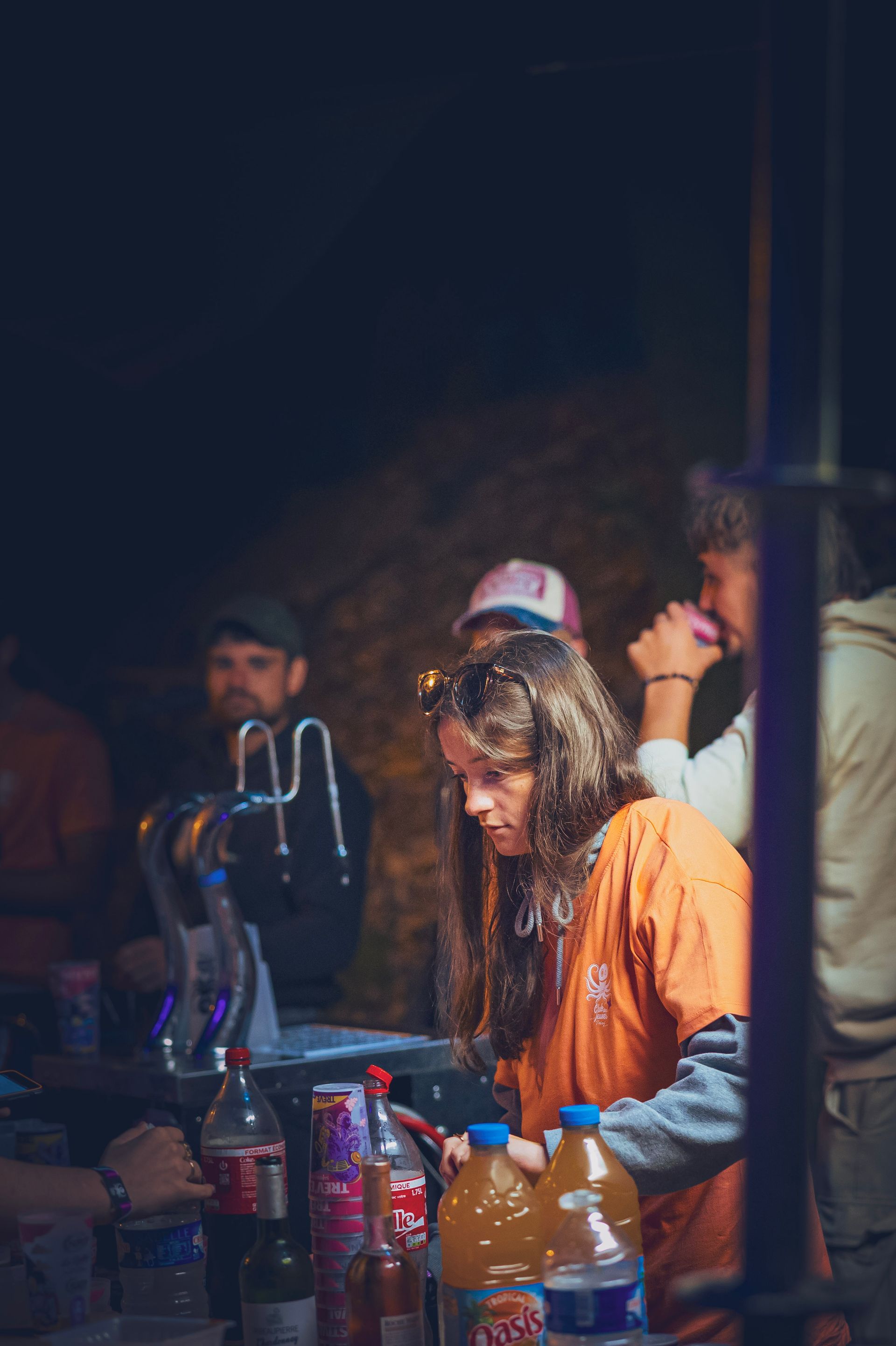 Une femme en chemise orange est assise à une table extérieure faiblement éclairée, avec des boissons et un narguilé, entourée de gens.