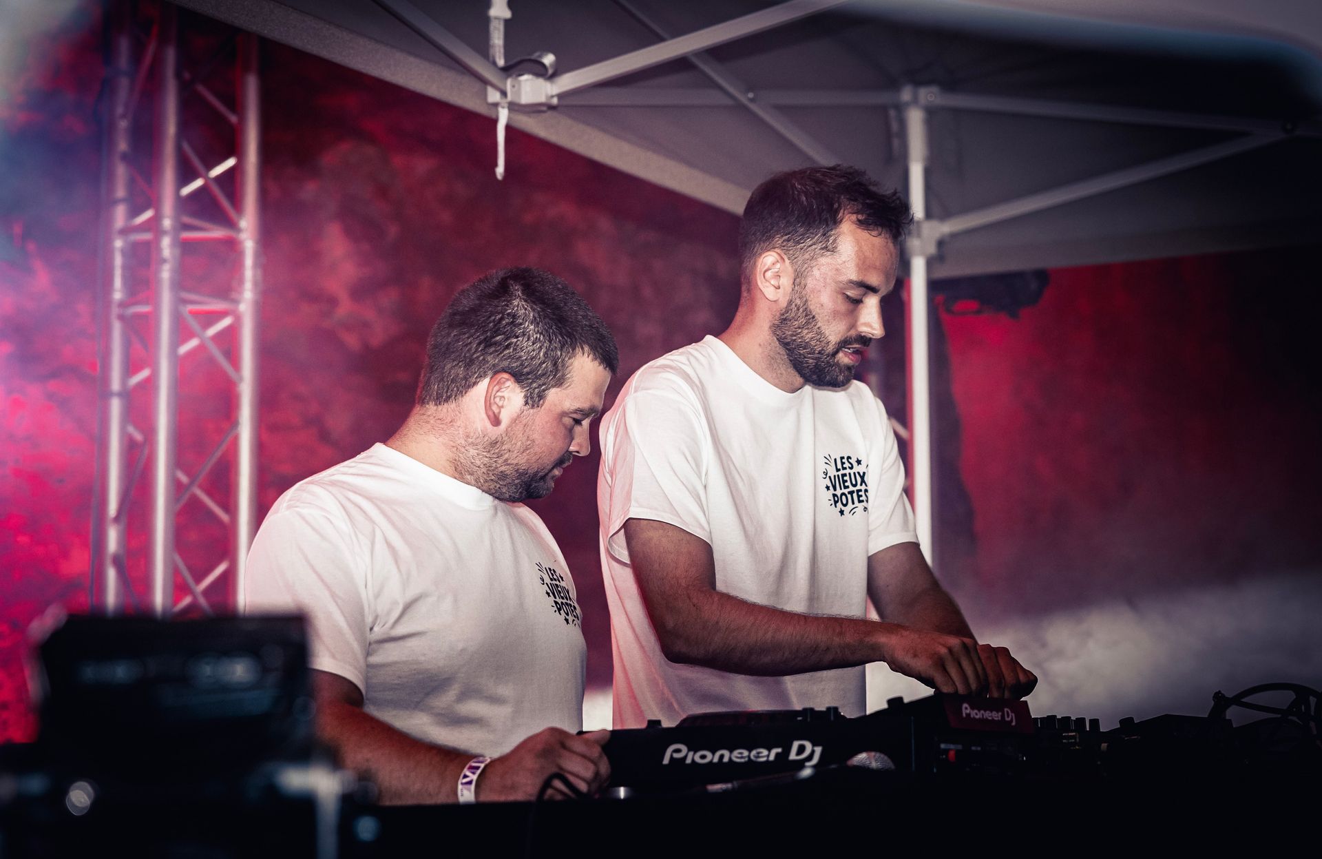 Deux DJ en chemises blanches mixant de la musique sous des lumières de scène roses lors d'un événement en plein air