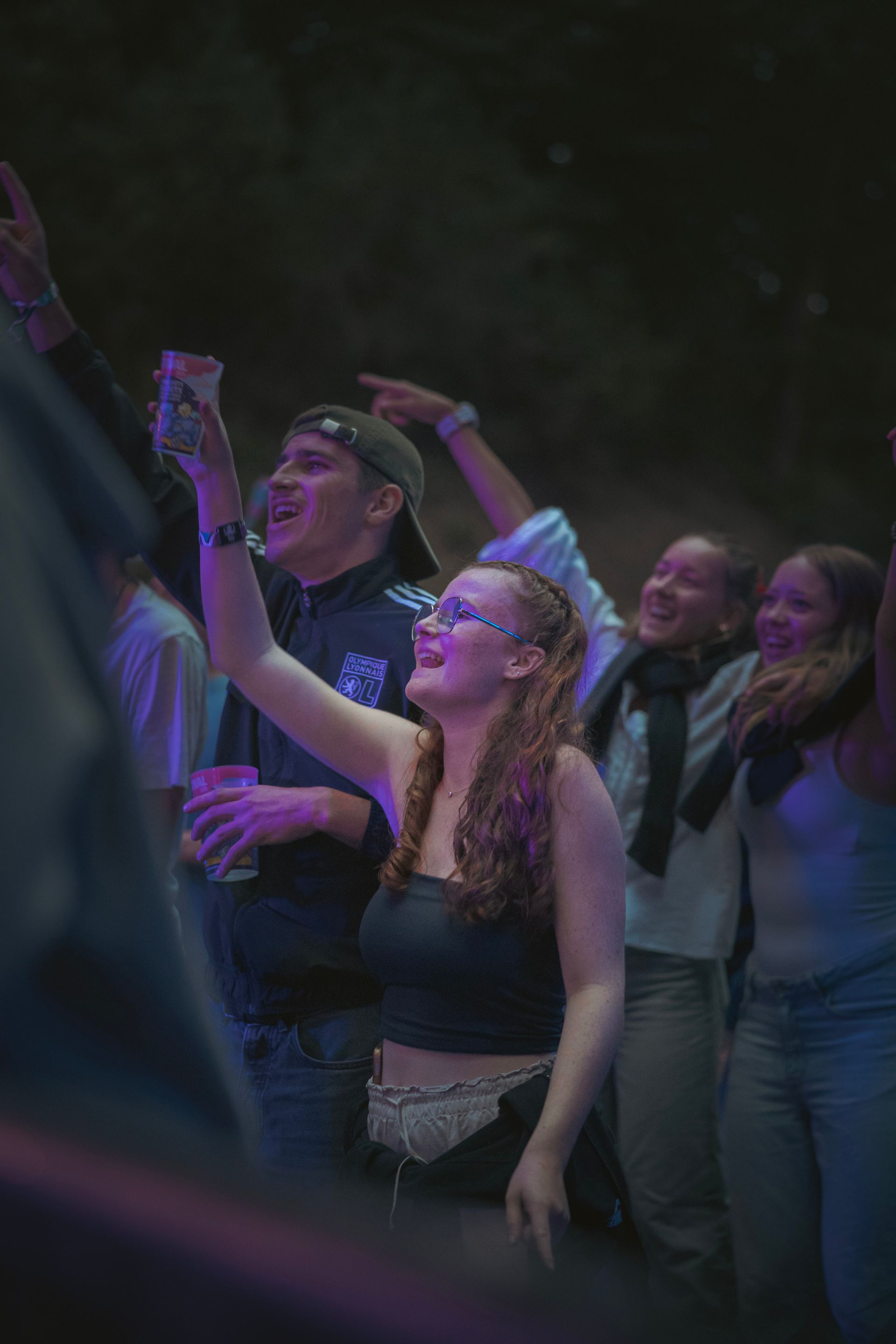 Foule dansant à un concert, une femme à lunettes levant le bras au premier plan