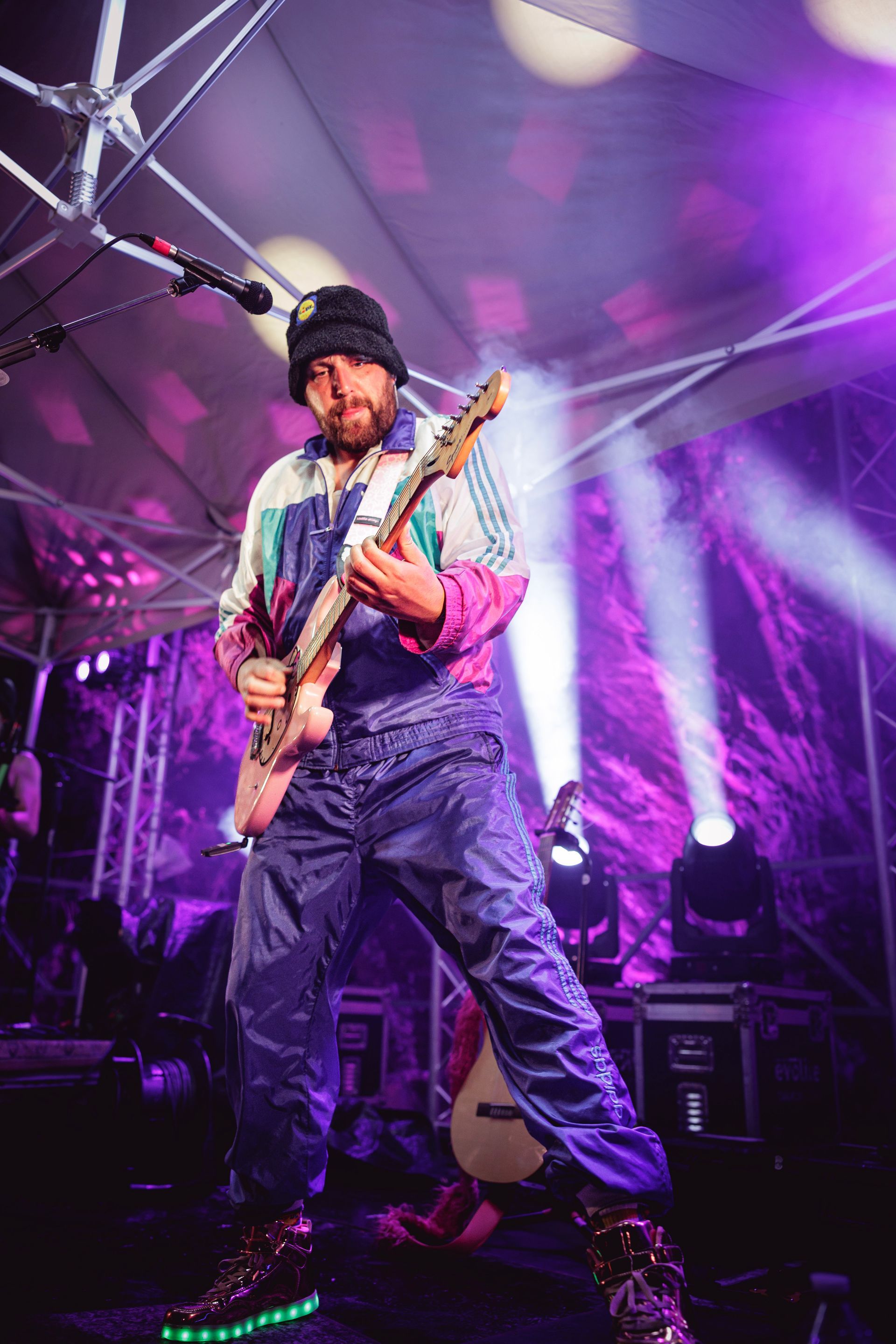 Un musicien, coiffé d'un bonnet et vêtu d'un survêtement coloré, joue de la guitare sous des lumières et des projecteurs violets.