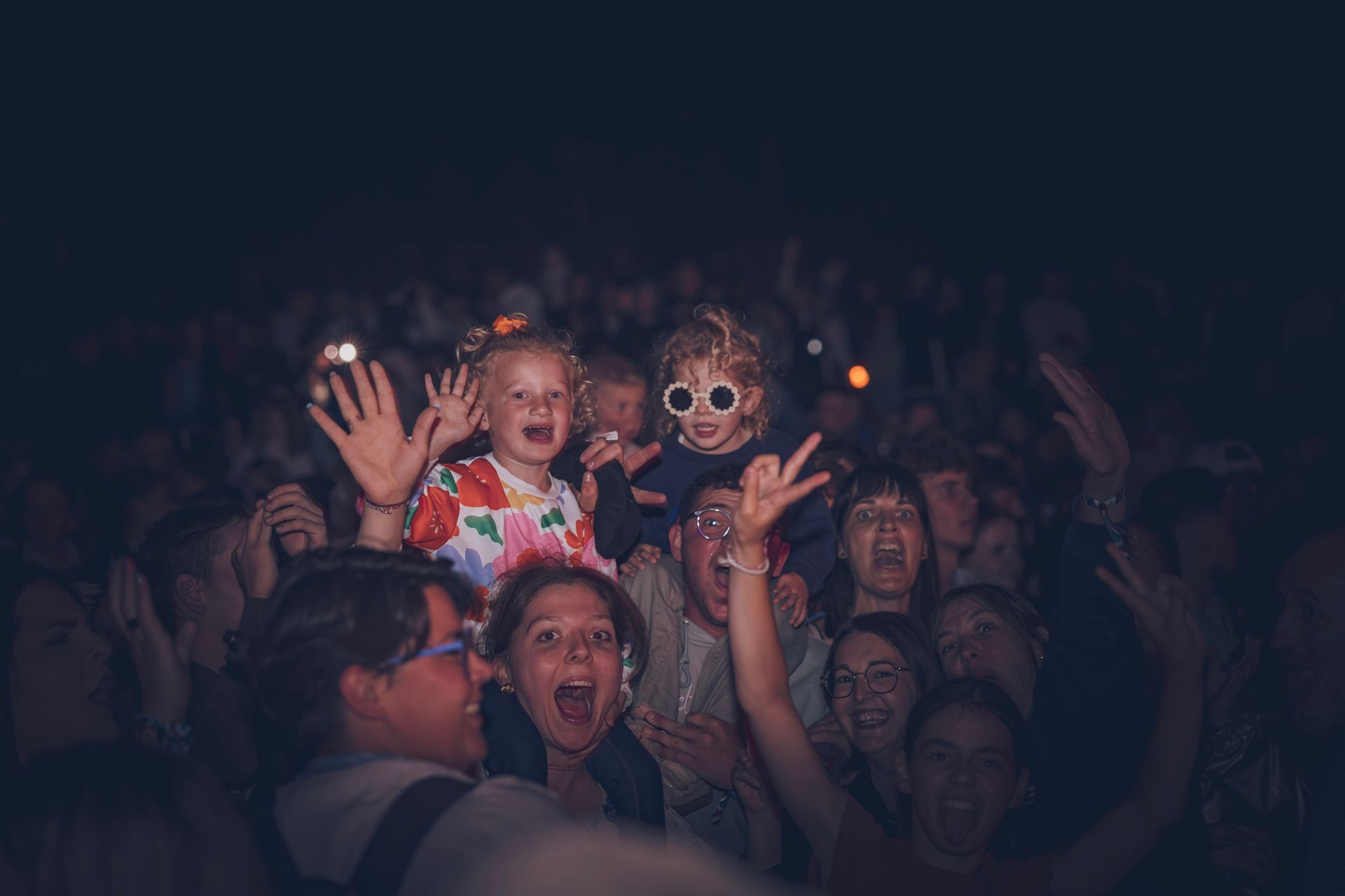 Foule à un concert, deux enfants sur les épaules d'adultes qui font coucou et sourient sous la faible lumière de la scène