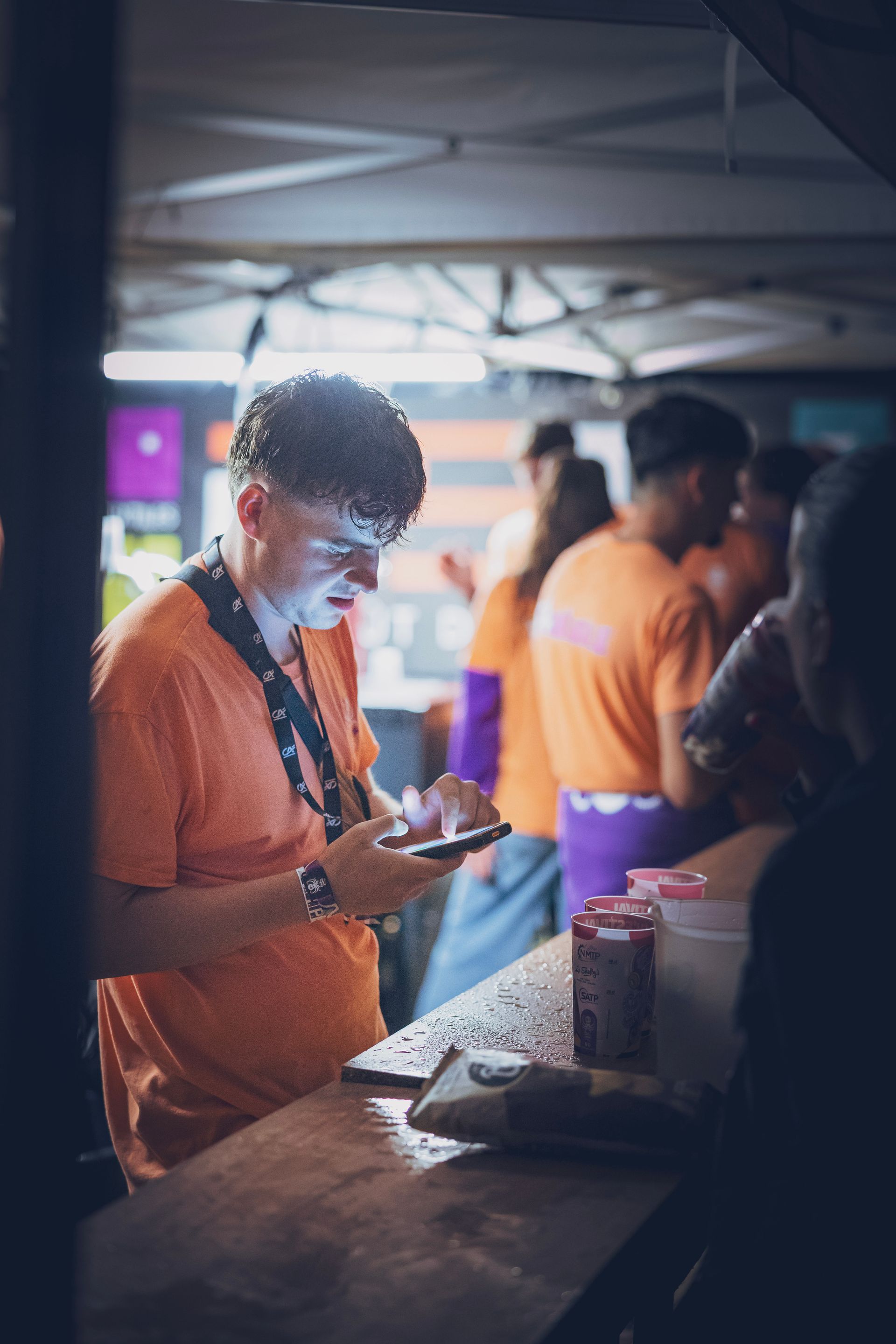 Une personne vêtue d'un t-shirt orange utilise un téléphone dans un stand extérieur aménagé sous un auvent.