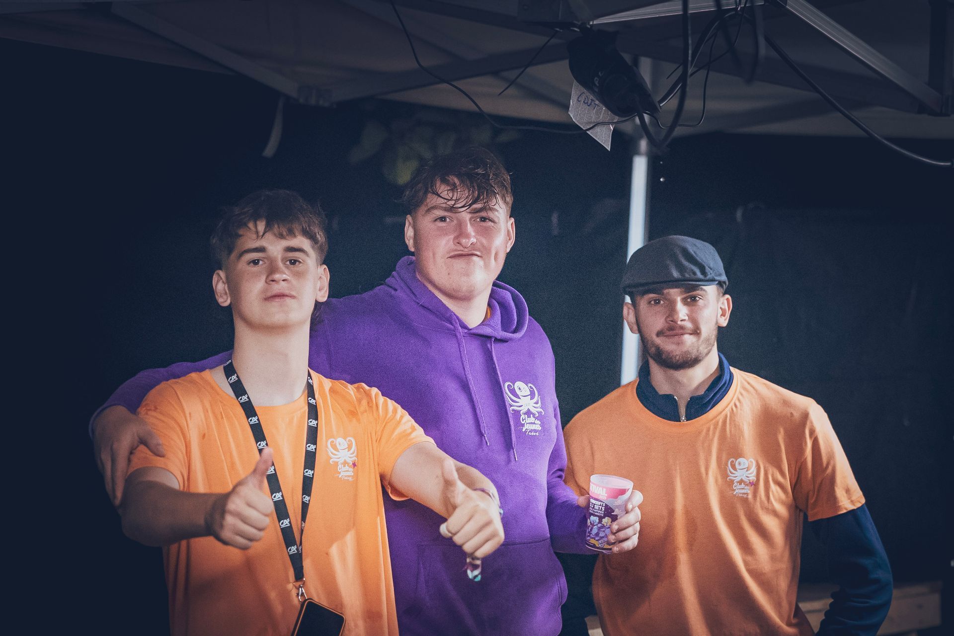 Trois hommes vêtus de chemises orange et violettes posent sous une tente, tenant des boissons et souriant.