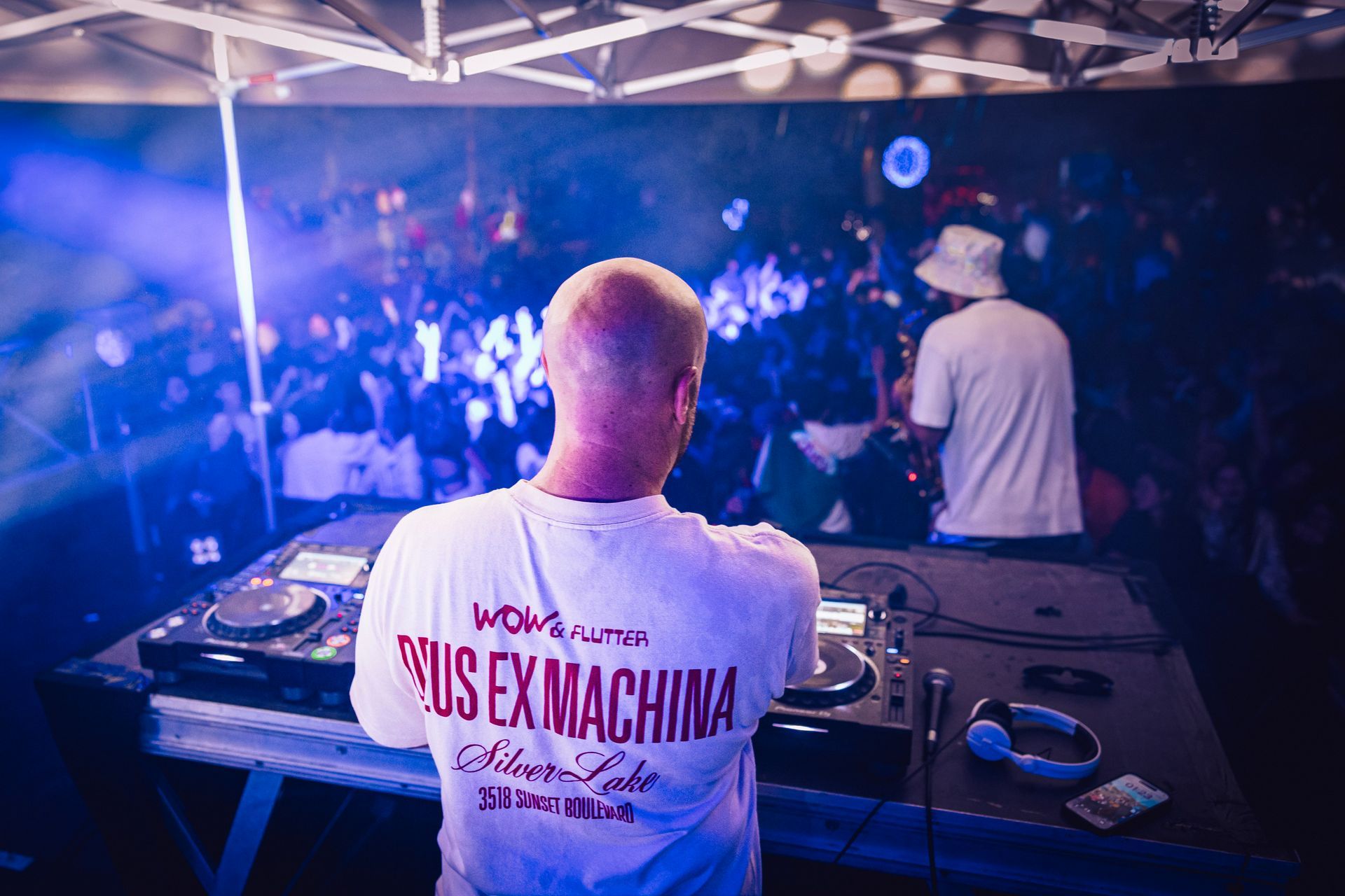 DJ aux platines, face à la foule sous des projecteurs bleu-violet, portant un t-shirt blanc « DEUS EX MACHINA »