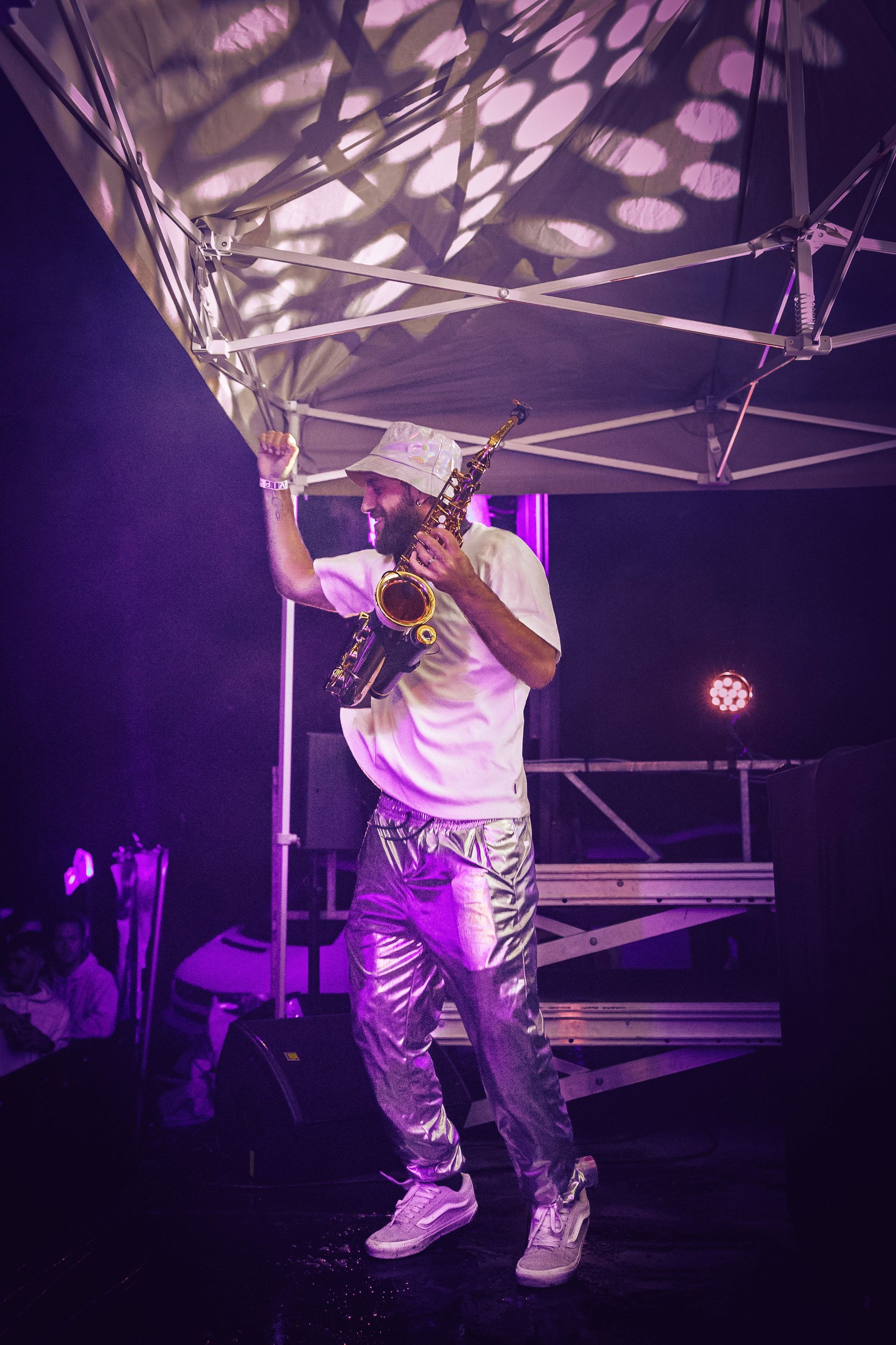 Un chanteur se produit sur scène sous des lumières violettes, vêtu d'une chemise blanche et d'un pantalon argenté, tenant un microphone.