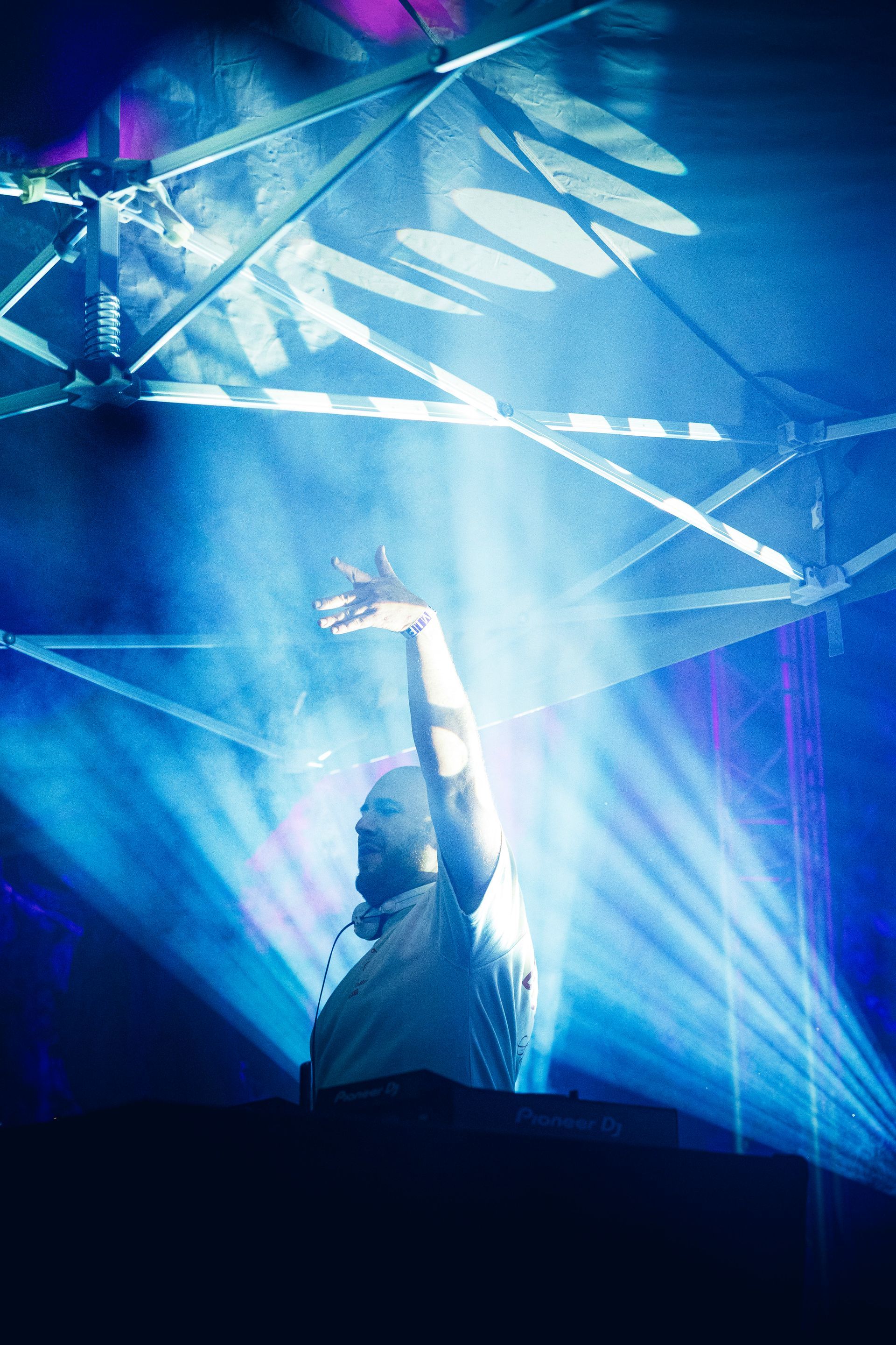 Un DJ lève un bras sur une scène éclairée en bleu, avec des faisceaux lumineux et des projections géométriques en arrière-plan.