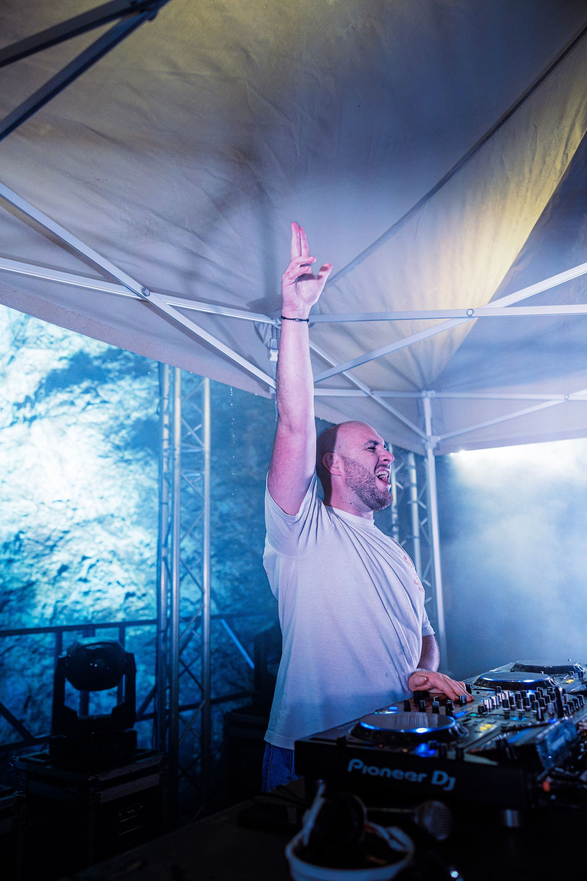 Un DJ en chemise blanche lève un bras sous un auvent lors d'un événement en plein air dans une atmosphère enfumée.