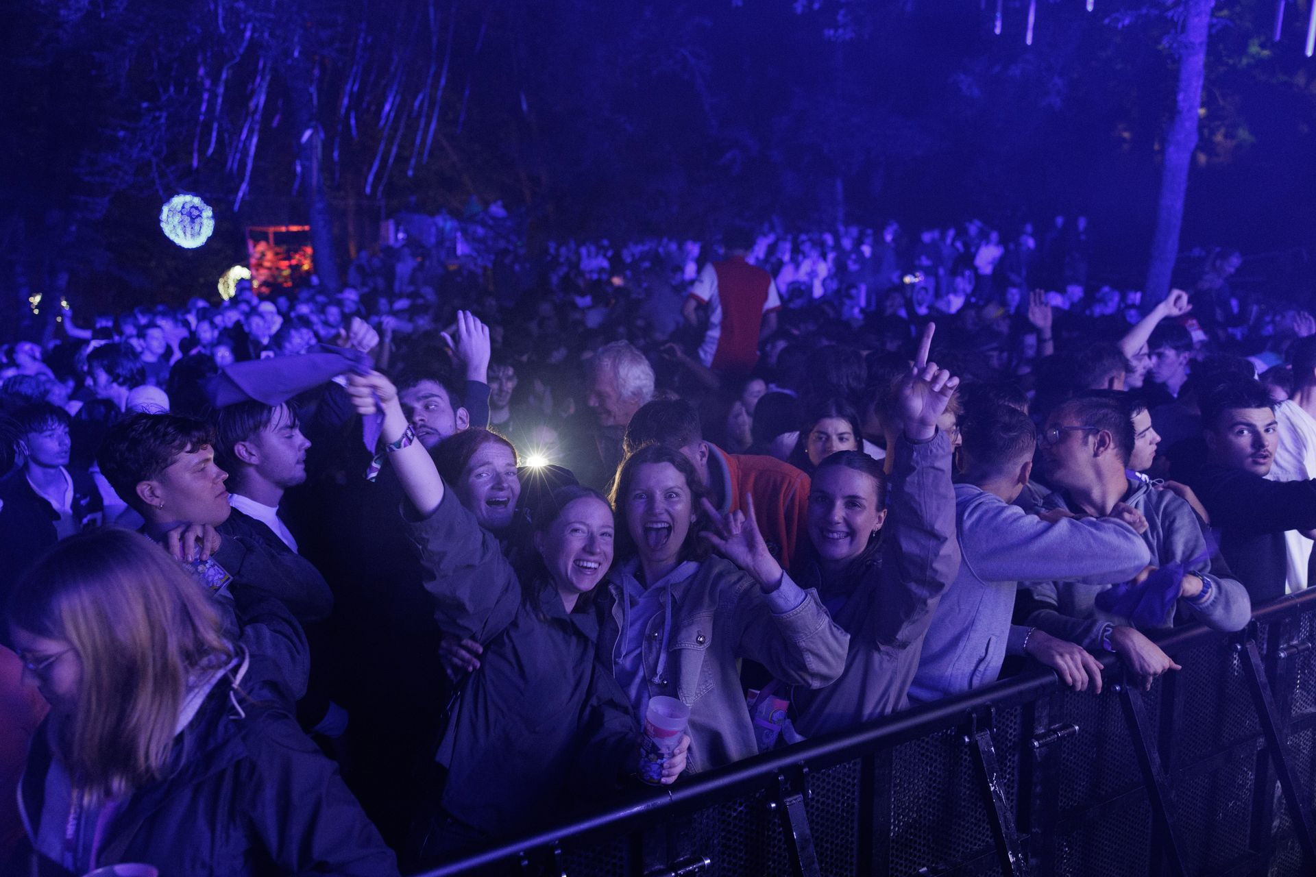 La foule acclame un concert nocturne en plein air, baignée de lumières bleu-violet, certains brandissant des téléphones.