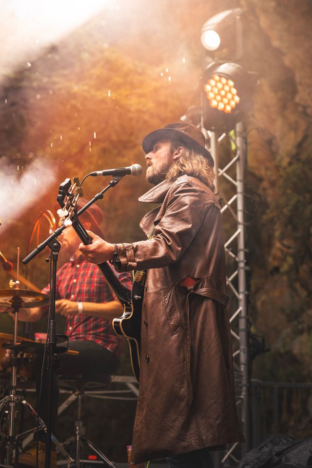 Un musicien en manteau et chapeau sombres chante dans un microphone sous les lumières chaudes de la scène