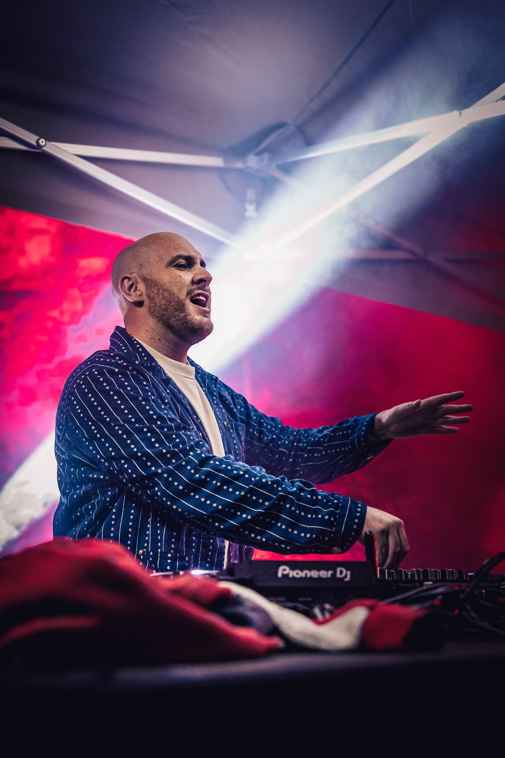 DJ se produisant sur scène sous des lumières roses et blanches, souriant avec un bras levé.