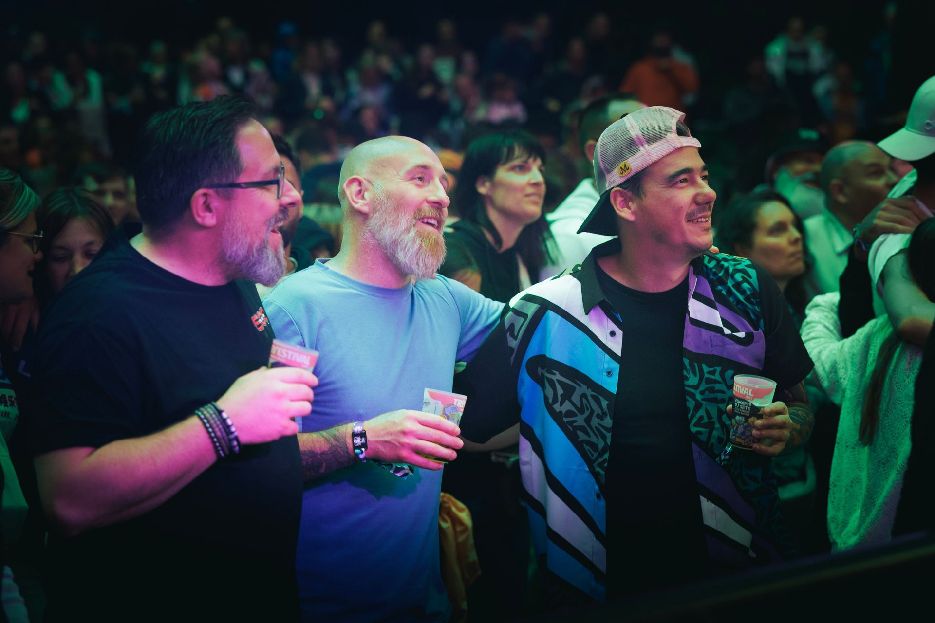 Trois hommes, verres à la main, sourient sous les lumières colorées de la scène lors d'un concert.
