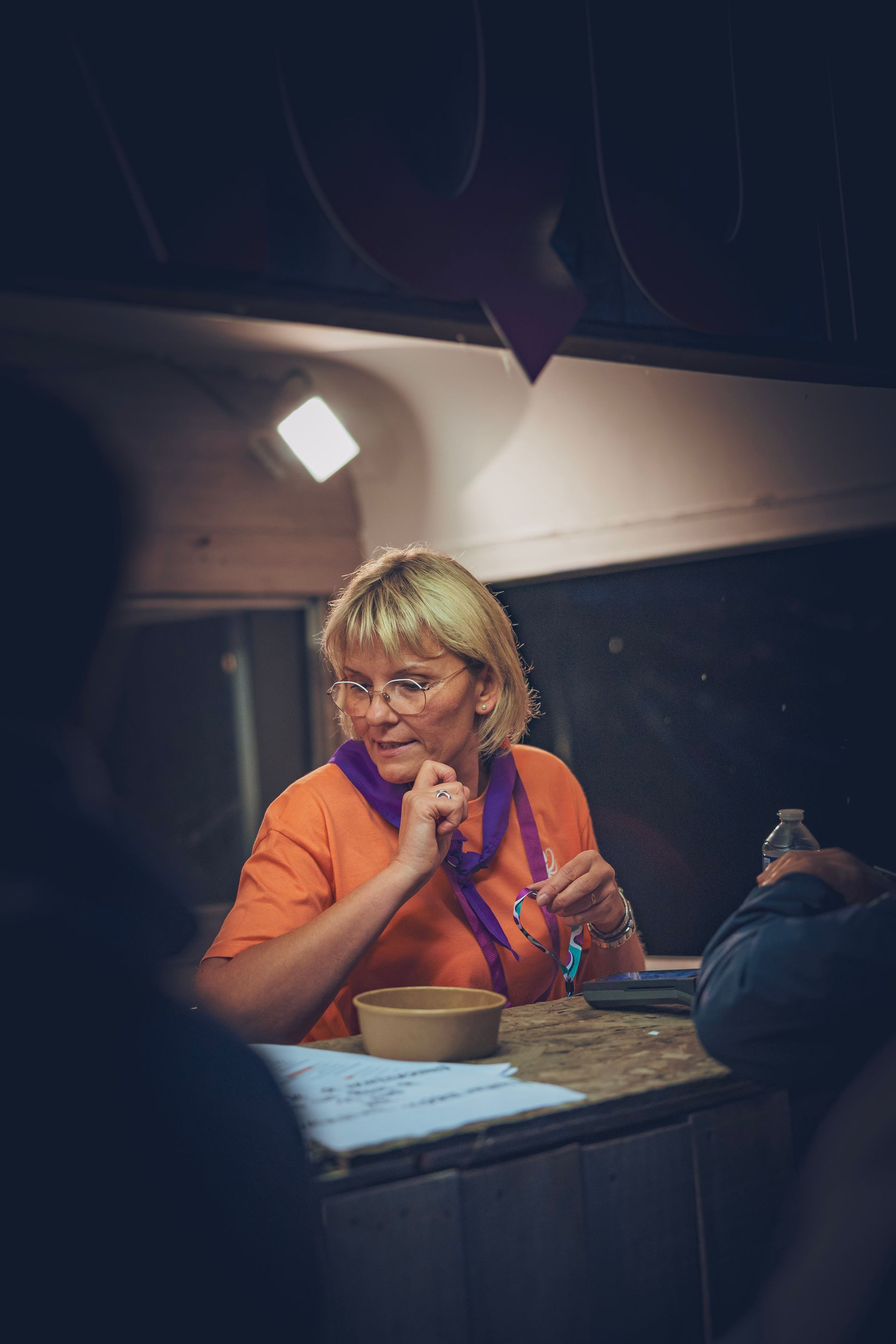 Une personne en chemise orange, assise à un comptoir, tient un téléphone sous un éclairage intérieur chaleureux.