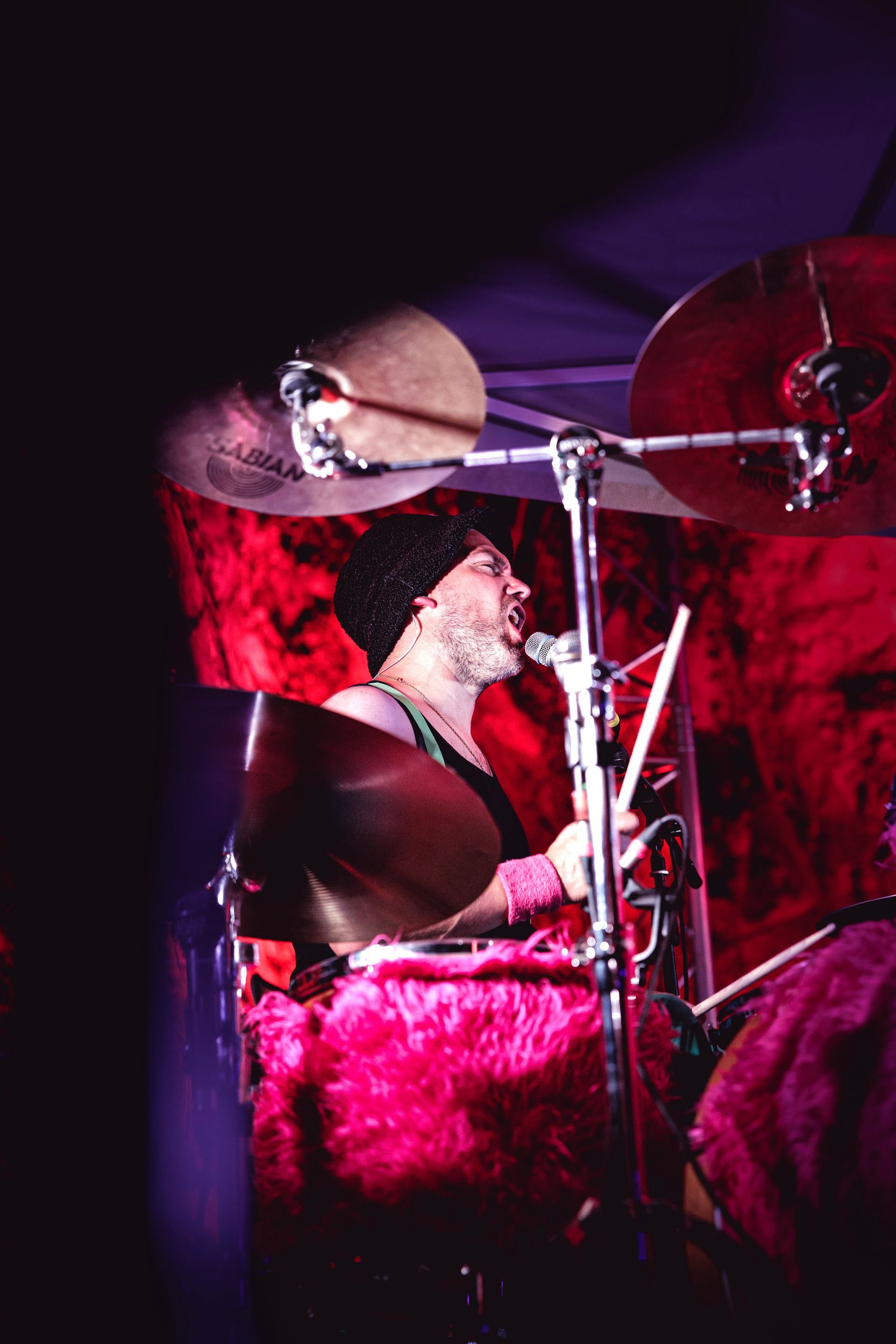 Un batteur se produit sur scène avec une batterie et des cymbales éclairées en rouge sous une lumière violette.