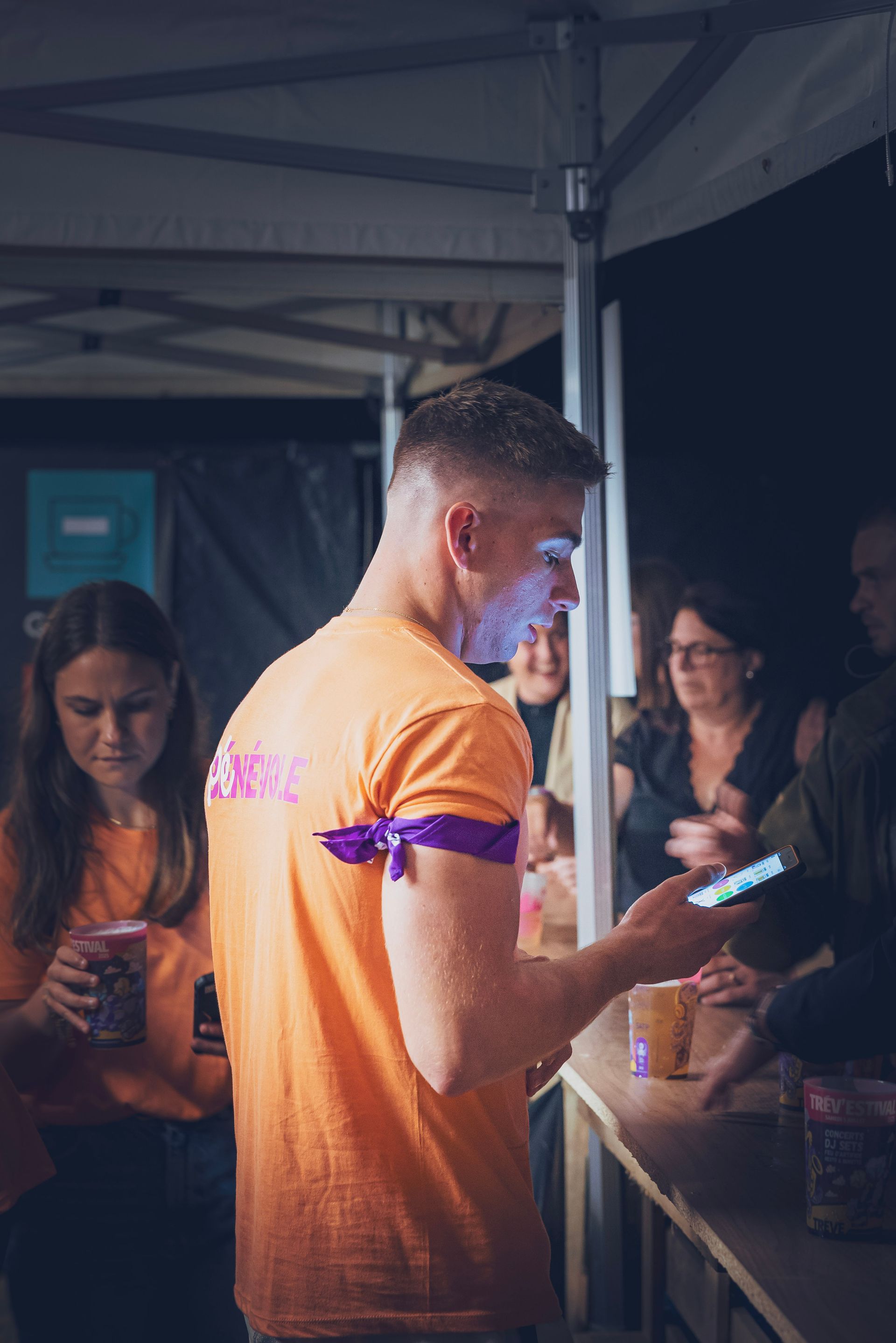 Une personne en chemise orange reçoit un bâton lumineux à un stand nocturne sous une tente