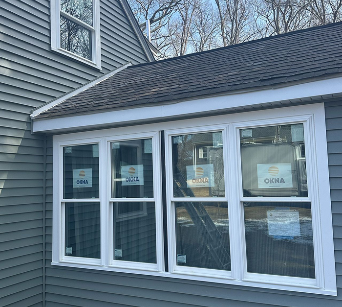 House exterior with white-framed windows and a dark shingled roof, partially covered in protective wrapping.