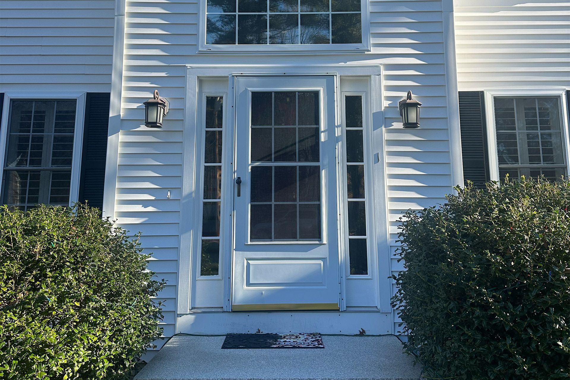 Front entry of a white house with a glass storm door, sidelights, black lanterns, and trimmed bushes